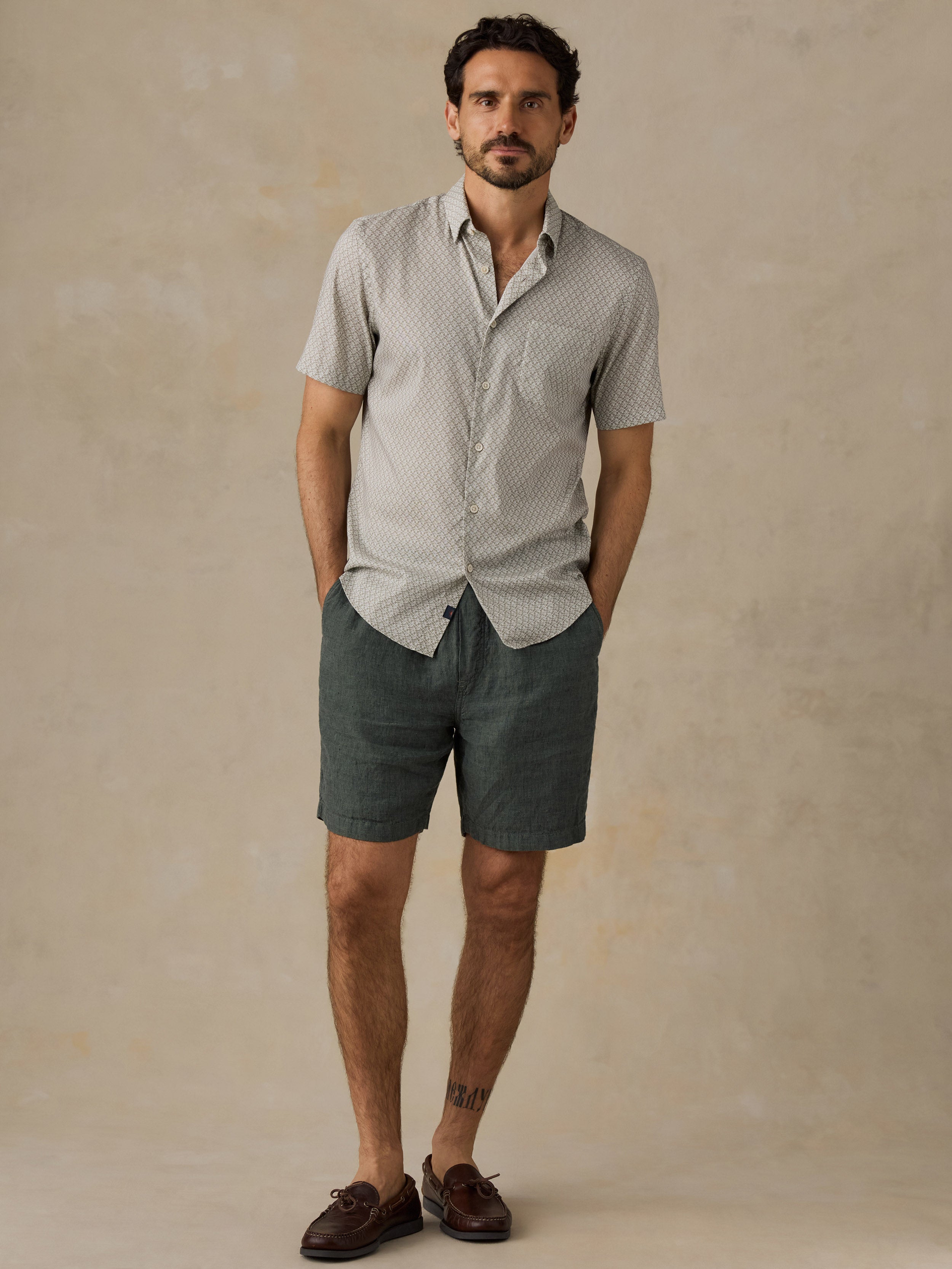 A man stands against a textured beige studio backdrop wearing a light green short-sleeve button-up shirt with a subtle geometric pattern, dark green shorts, and brown leather boat shoes.