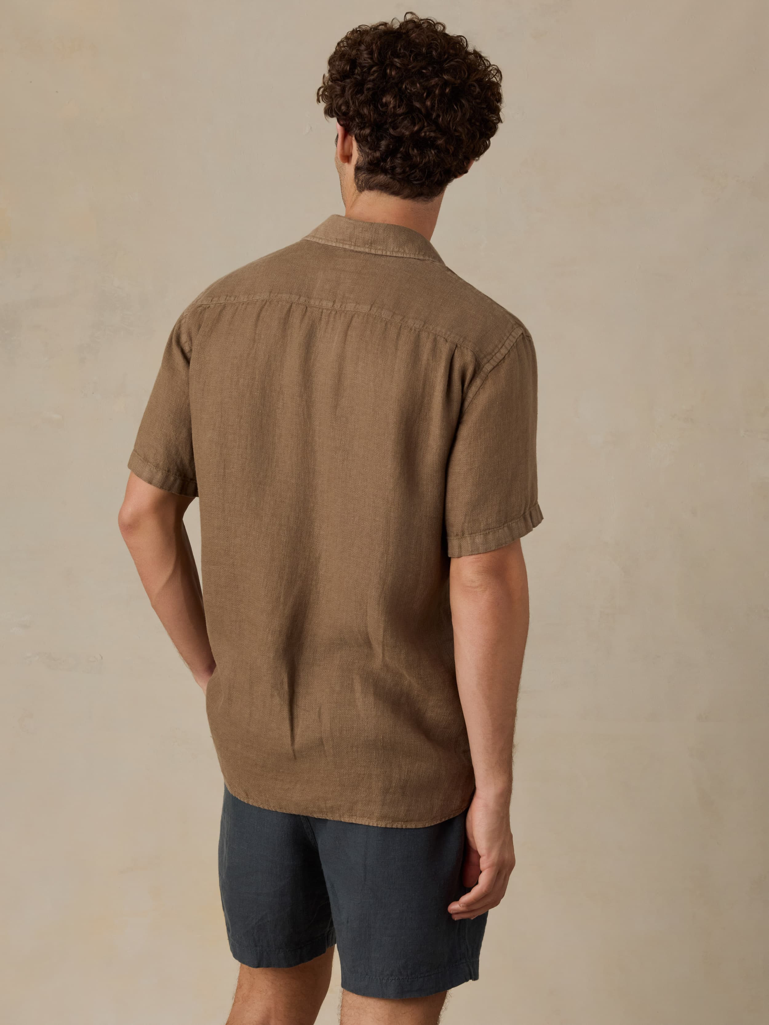 Man wearing a brown short-sleeve shirt and dark shorts against a beige background