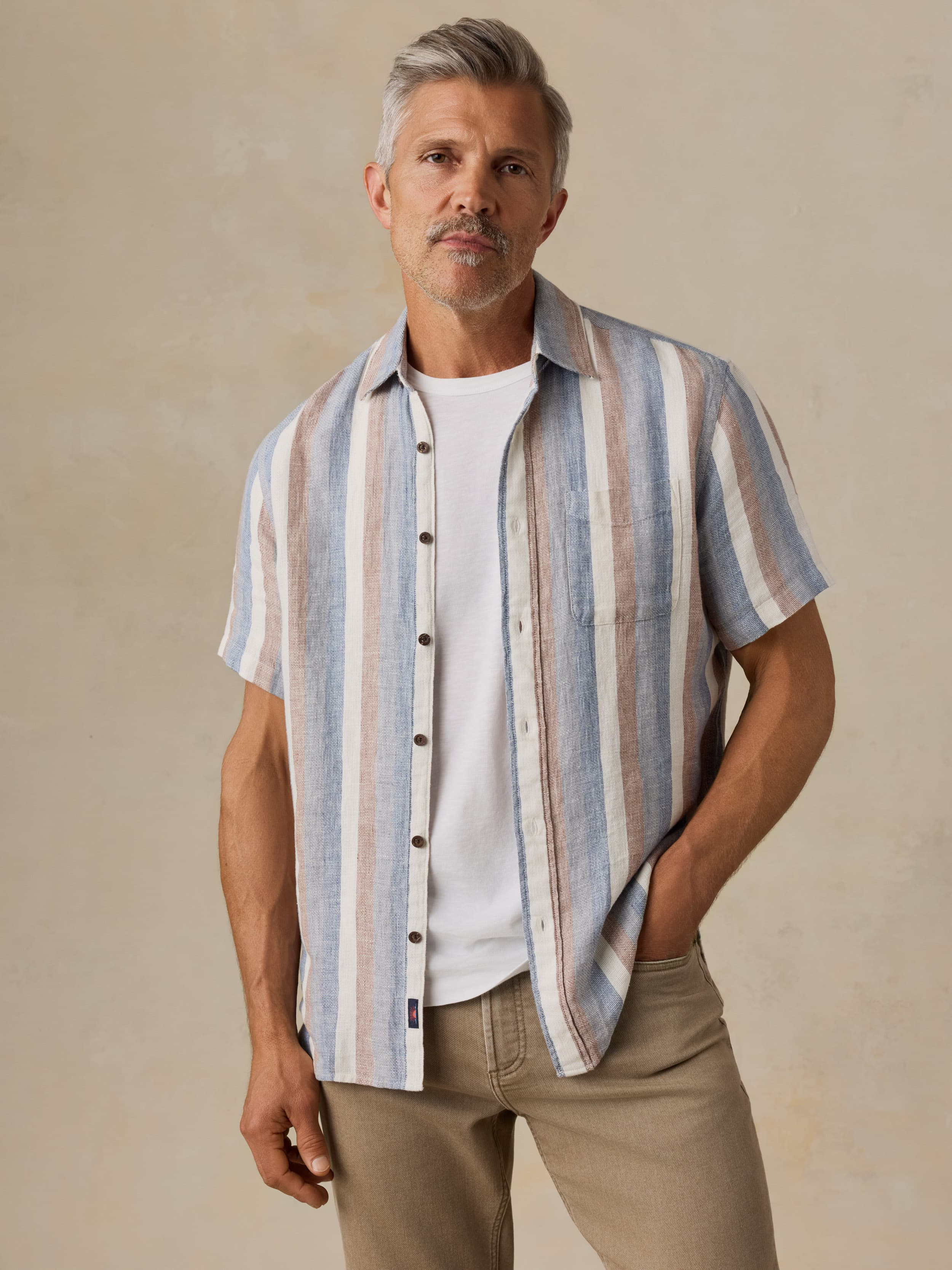 Man wearing a striped shirt over a white t-shirt with beige pants against a beige background