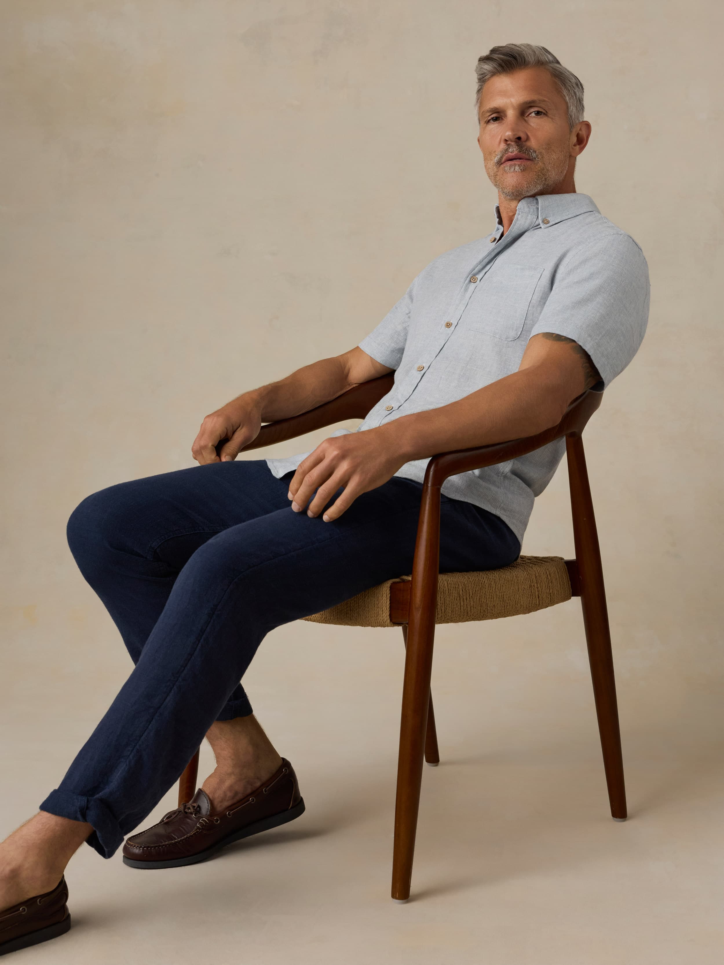 Man sitting in a chair wearing a light blue short-sleeve shirt and dark blue pants against a beige background