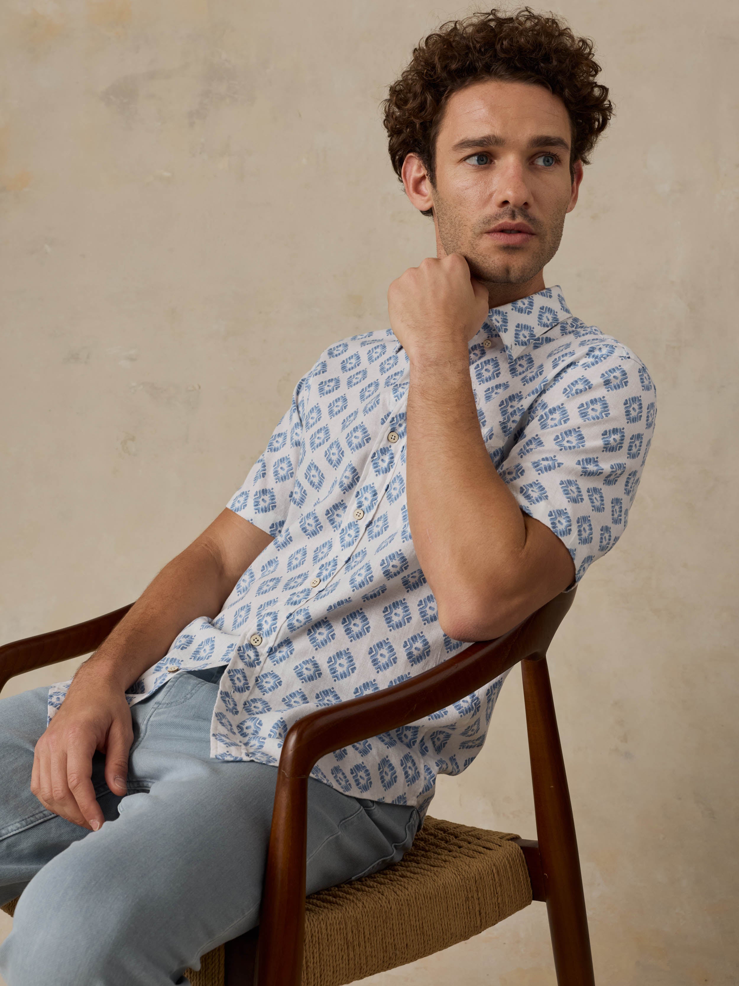 Man sitting on a wooden chair wearing a patterned blue and white shirt against a beige background