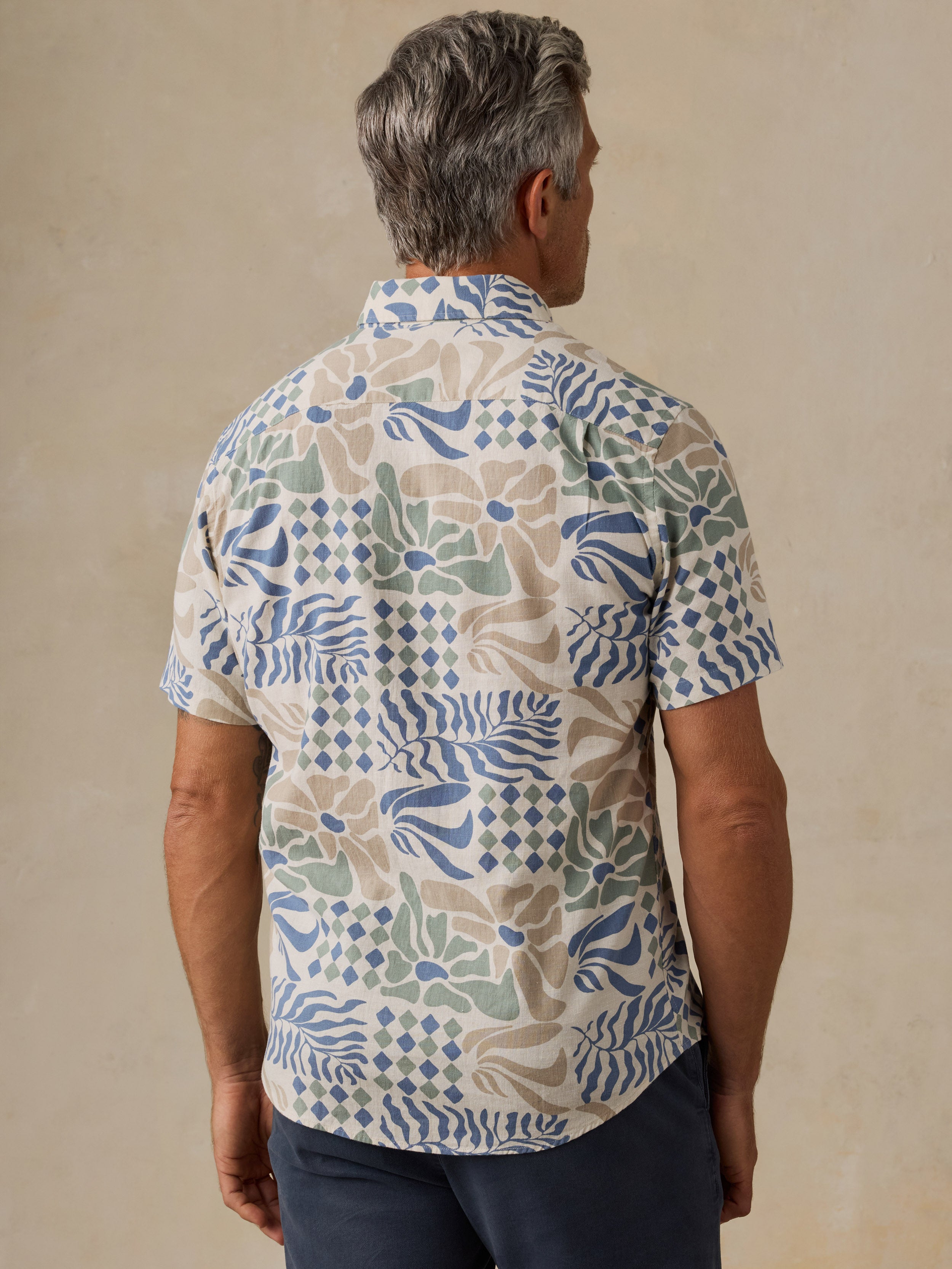 A man viewed from behind wearing a short-sleeve button-up shirt with a multicolor abstract botanical pattern in blue, green, and beige on a cream base, paired with navy pants, set against a neutral beige background.