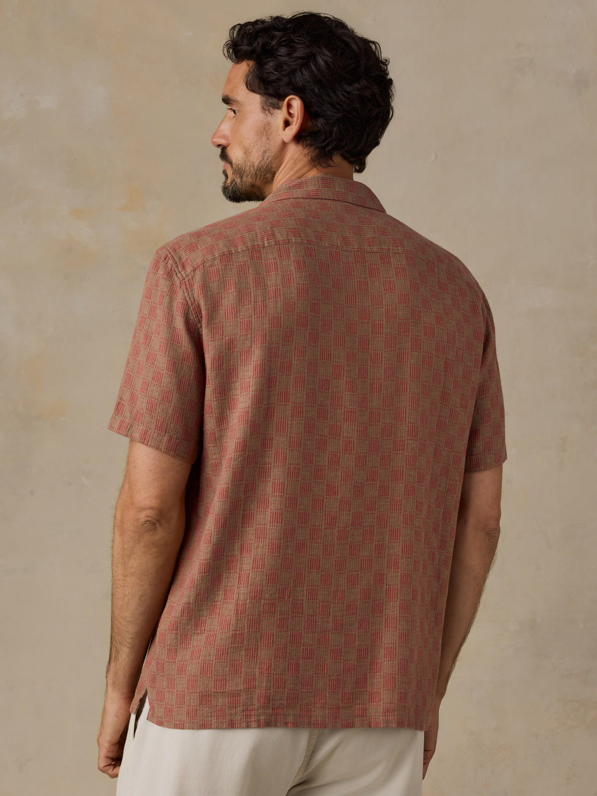 A man wearing a short-sleeve button-up shirt with a muted reddish-brown and beige geometric pattern, shown from the back against a textured beige wall., image 4 of 7