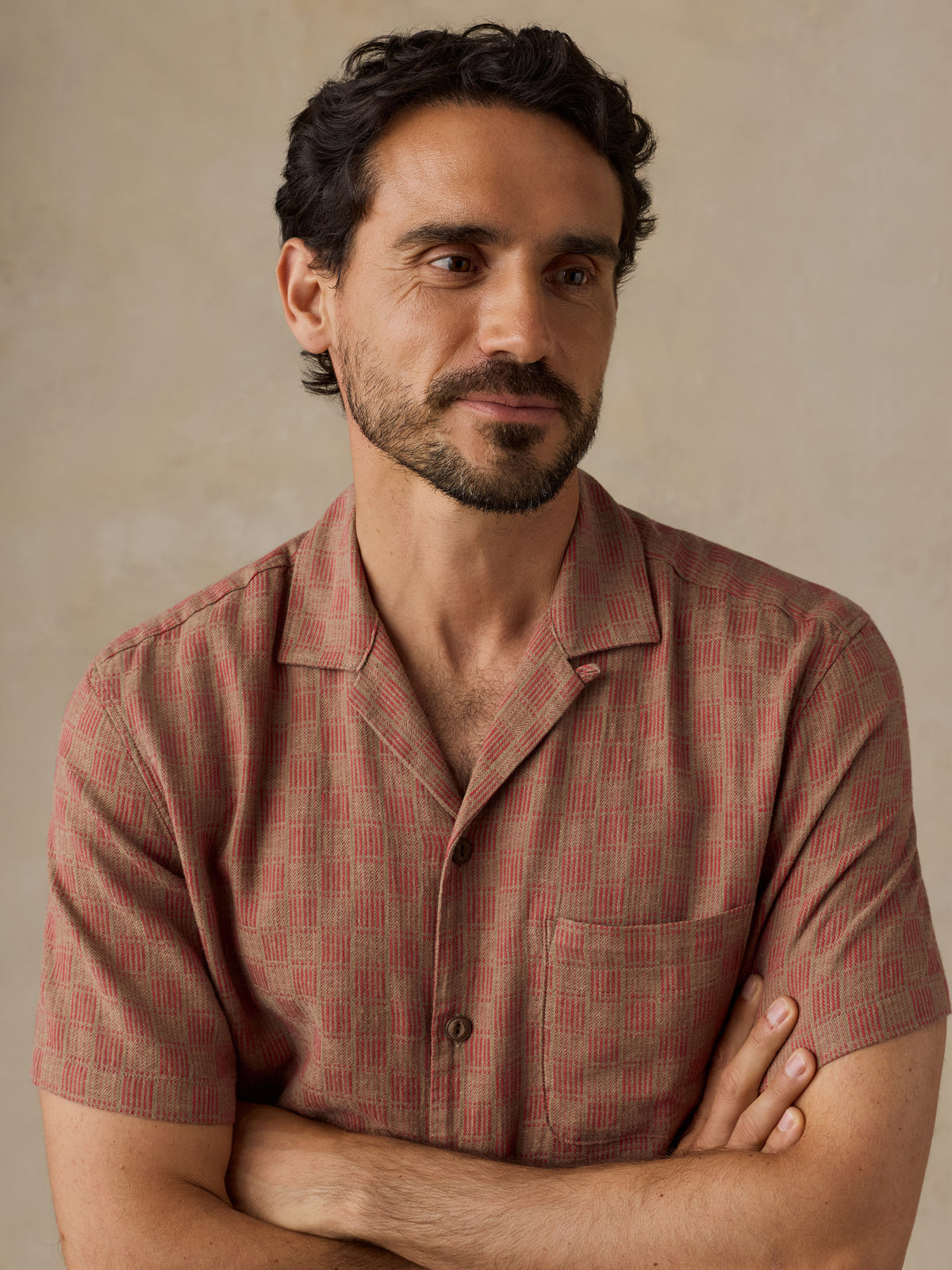 A man with dark curly hair and a beard wearing a short-sleeve button-up shirt with a red and beige geometric pattern, posed with arms crossed against a neutral beige background., image 3 of 7