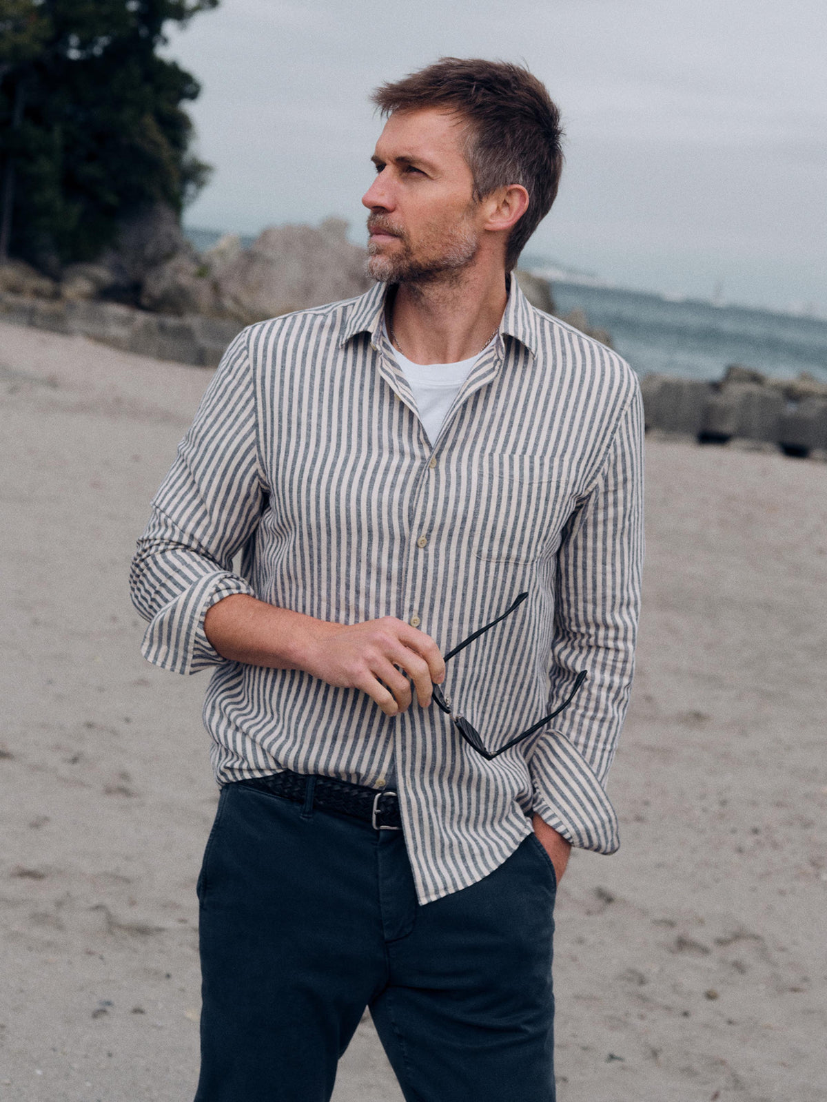 A man standing on a sandy beach wearing a vertically striped button-down shirt in beige and navy, dark trousers, and holding sunglasses., image 1 of 6