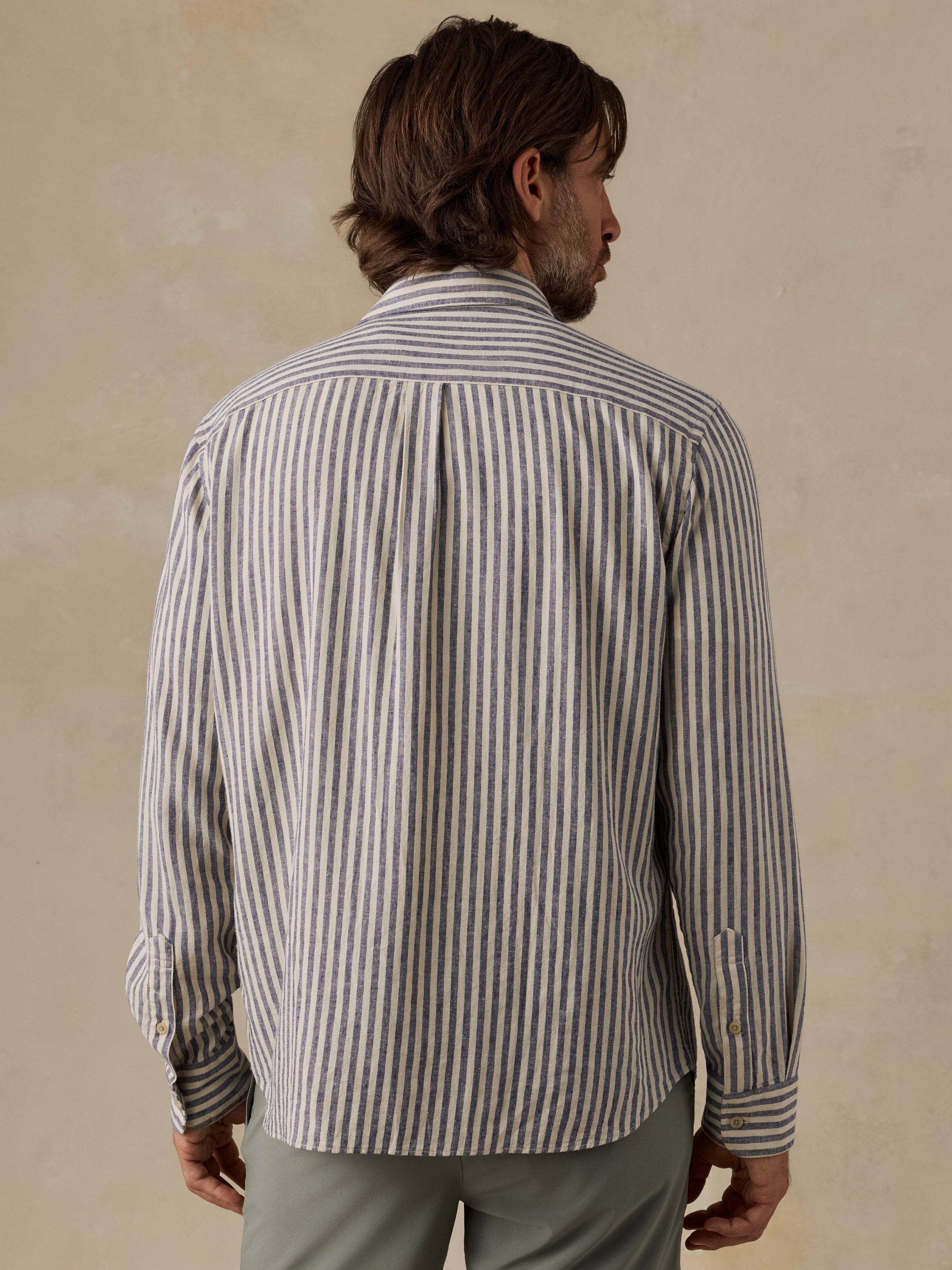 A man viewed from behind wearing a long-sleeve button-up shirt with vertical gray and white stripes, paired with muted green shorts, set against a textured beige background.