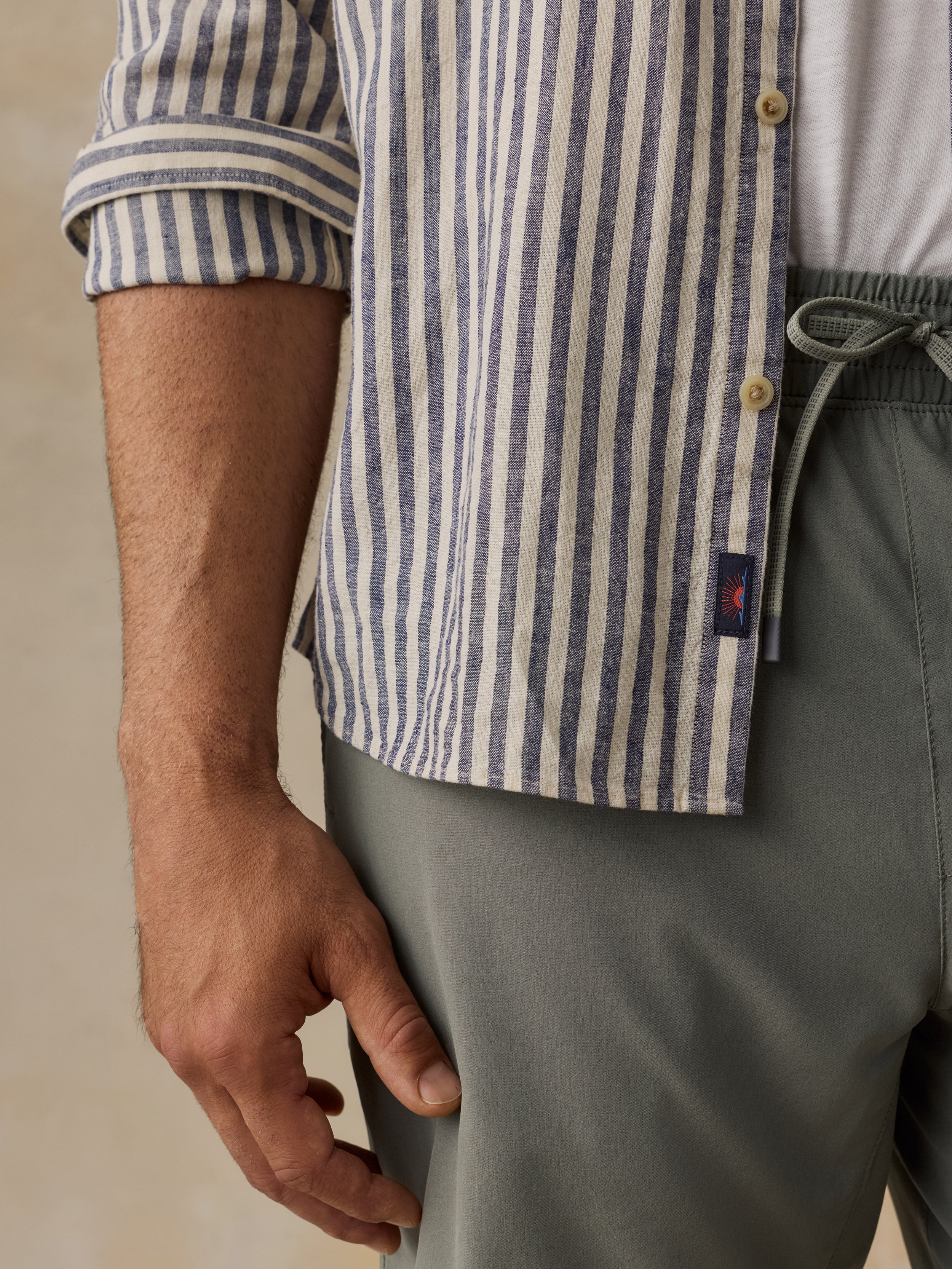 Close-up of a man wearing a blue and white vertically striped button-up shirt with rolled sleeves, paired with gray drawstring trousers.