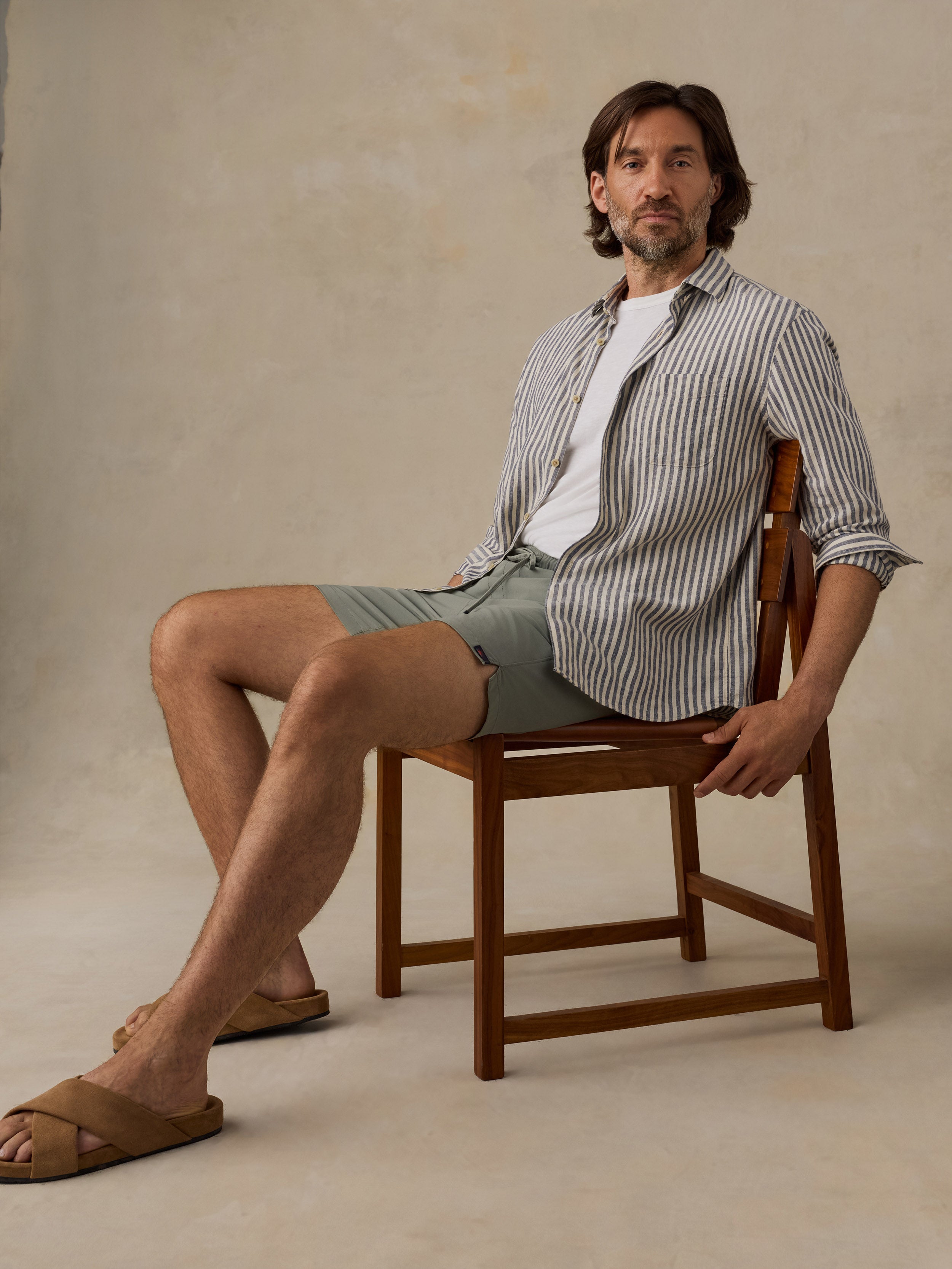 A man sits on a wooden chair wearing a white t-shirt, a striped button-down shirt, sage green shorts, and brown slide sandals against a textured beige studio backdrop.