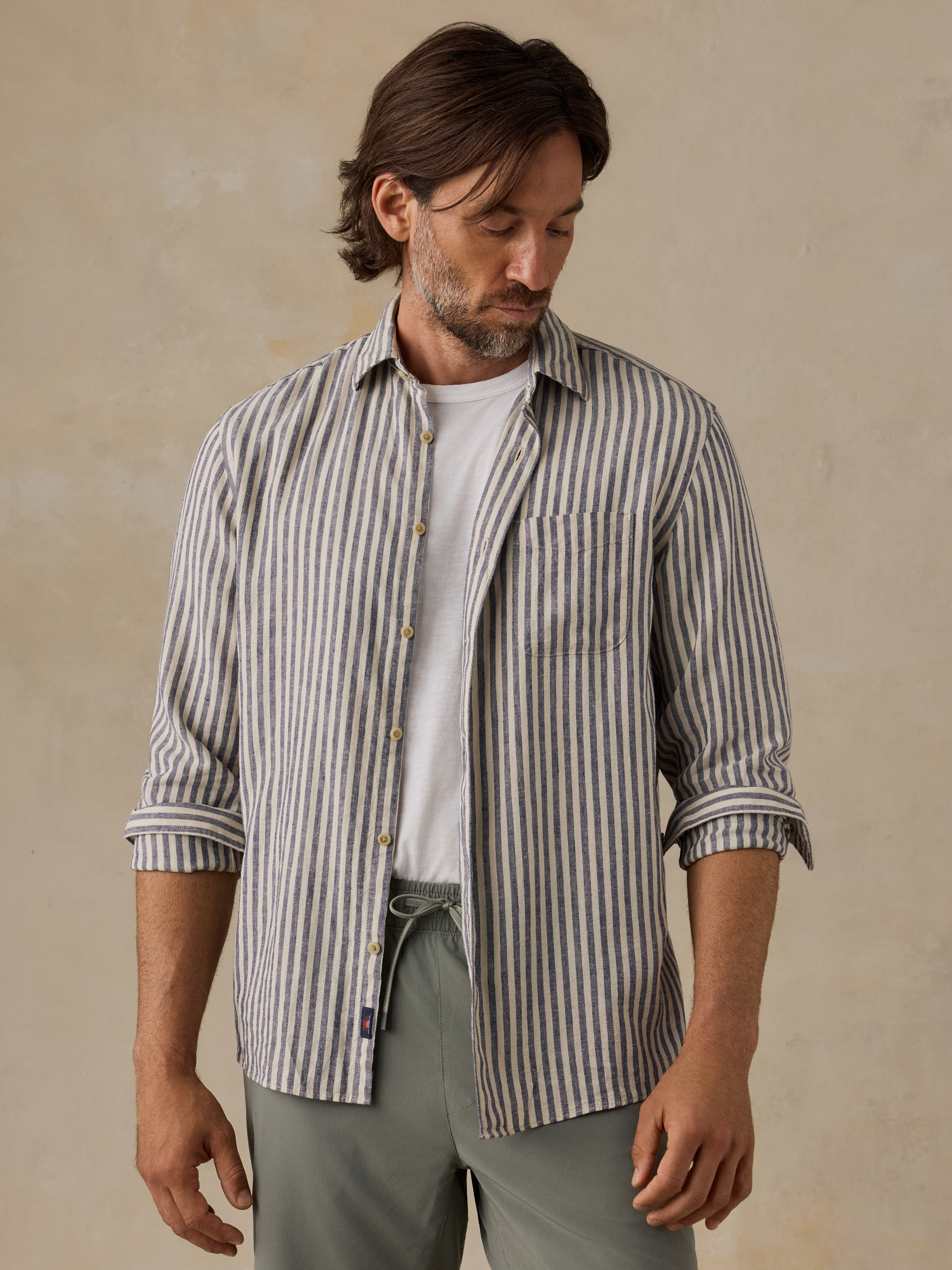 A man wearing a long-sleeve button-up shirt with vertical gray and white stripes over a white t-shirt, paired with olive green pants, set against a textured beige background.