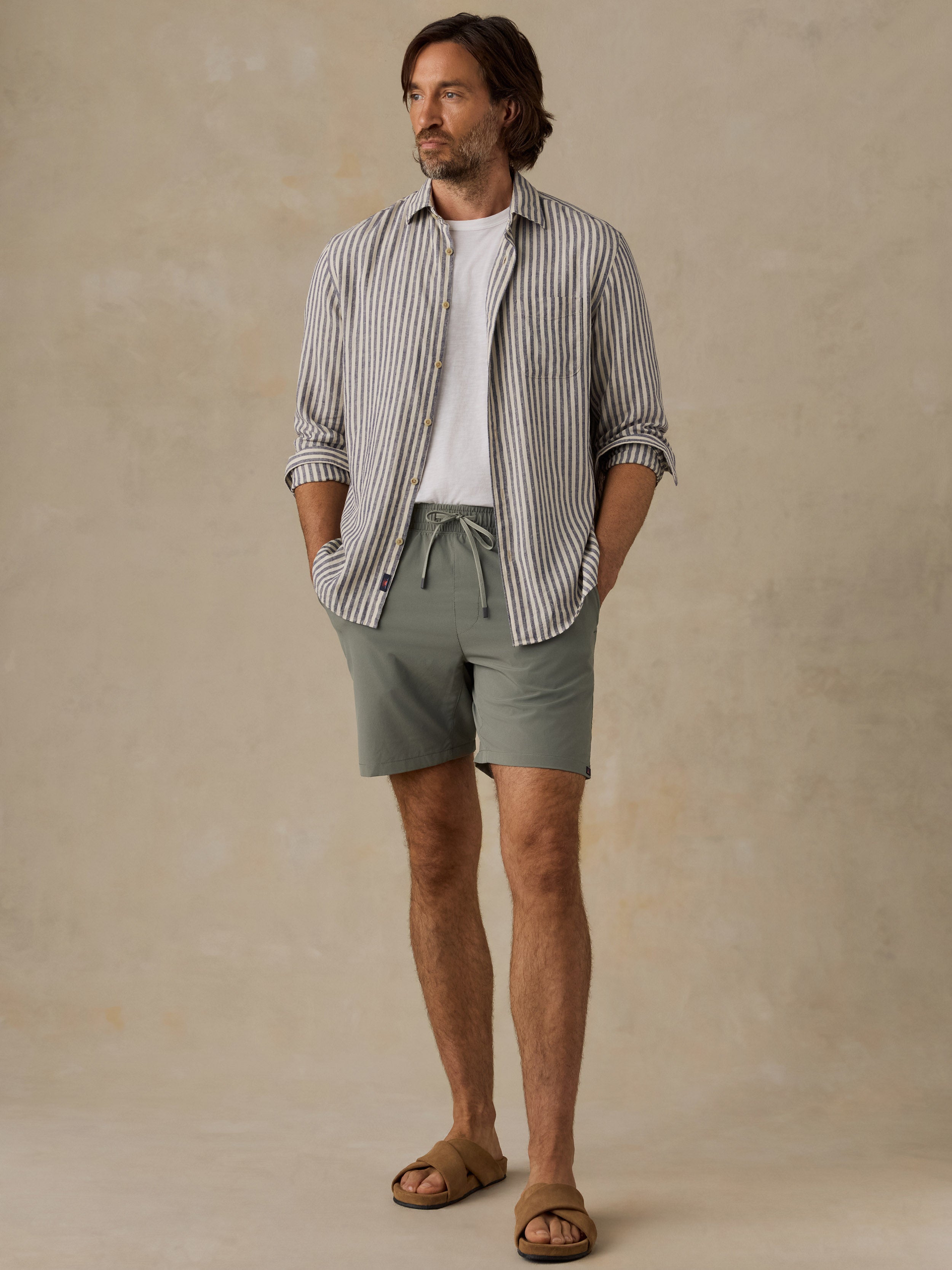 A man stands against a warm beige textured studio backdrop wearing a striped button-up shirt over a white tee, sage green drawstring shorts, and brown slide sandals.