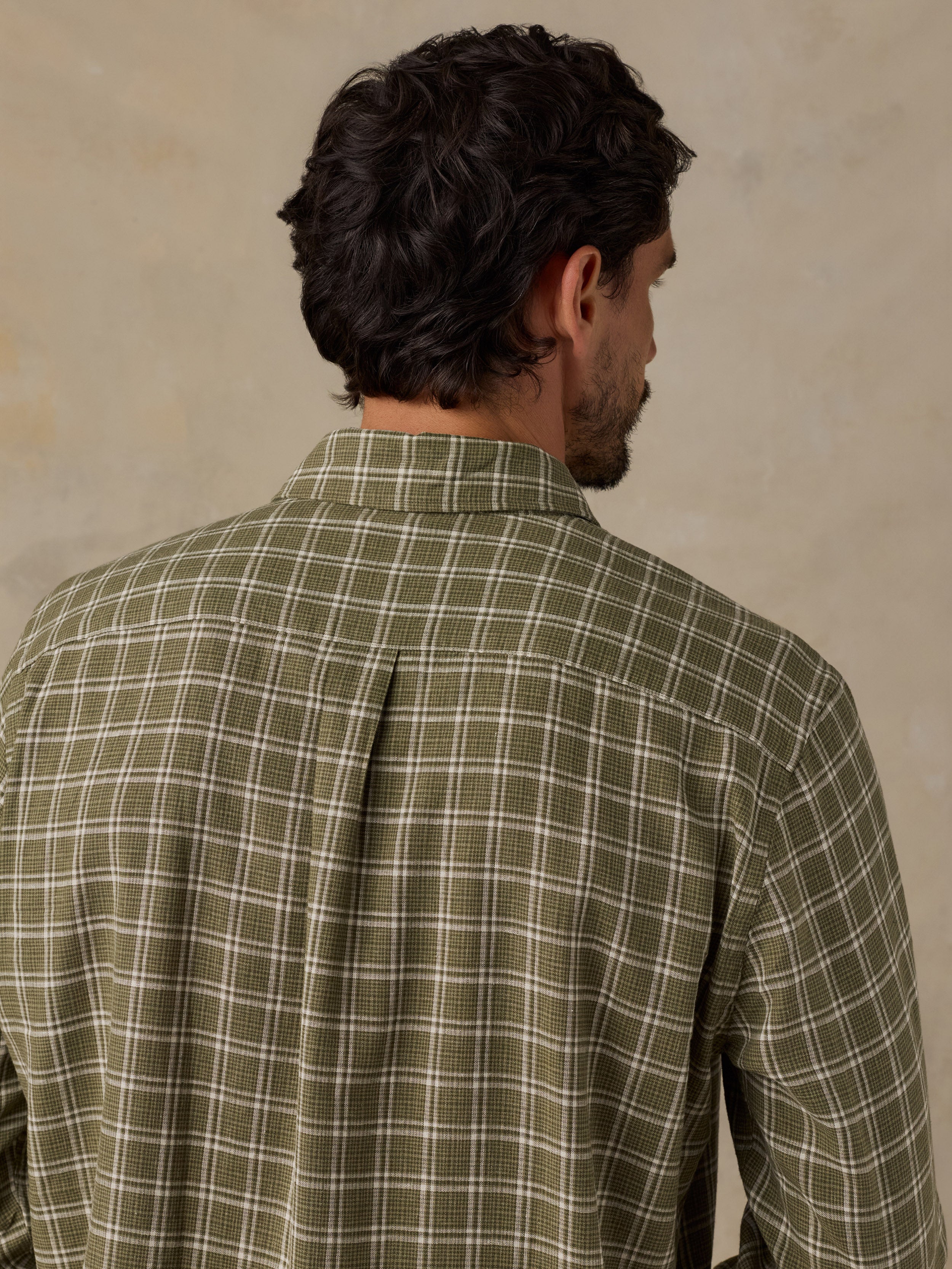 A man viewed from behind wearing a long-sleeve plaid button-down shirt in olive green and white, set against a textured beige background.