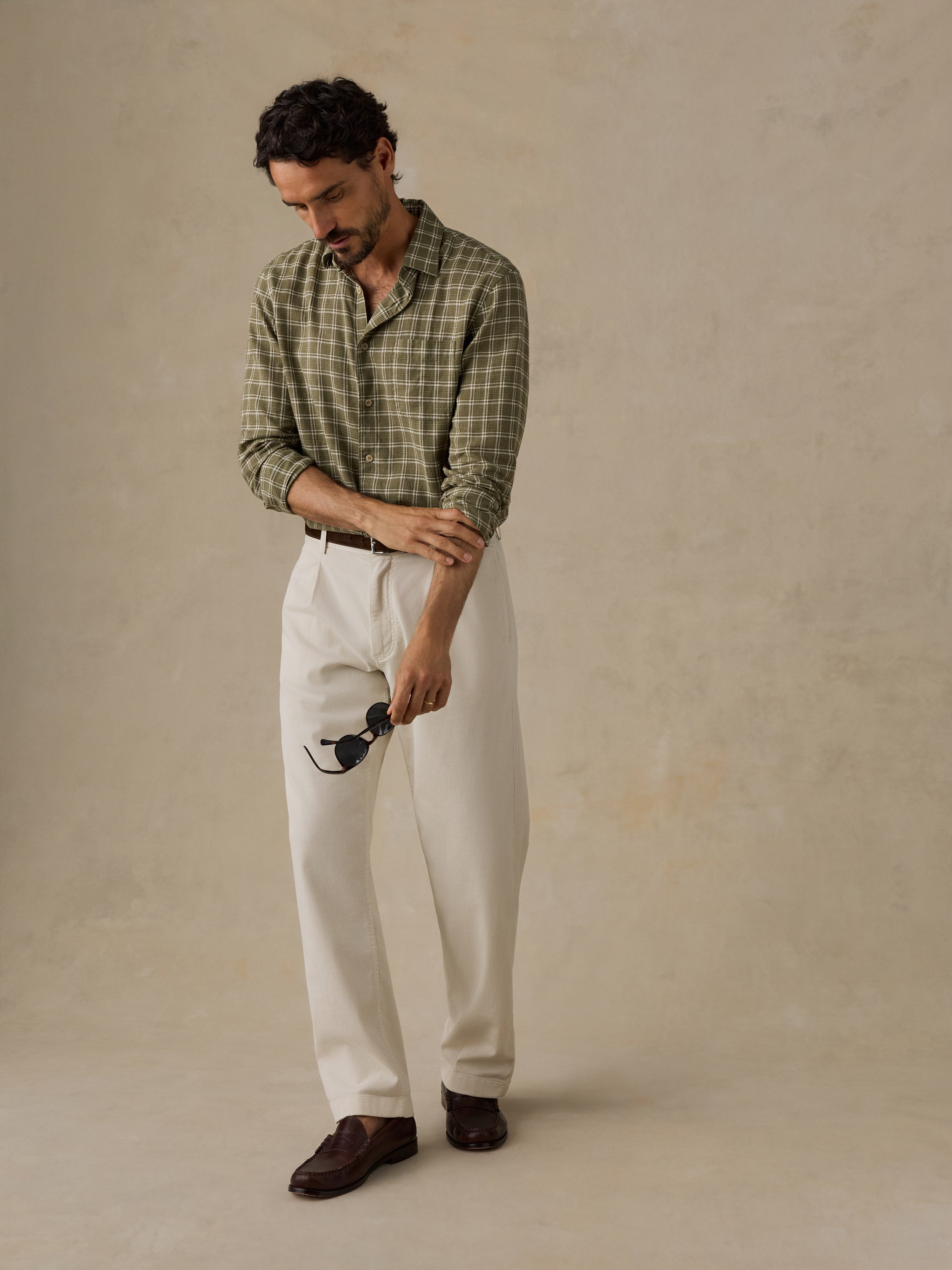 A man wearing a green and beige plaid button-down shirt, off-white trousers, and dark brown loafers, standing against a textured beige studio backdrop.