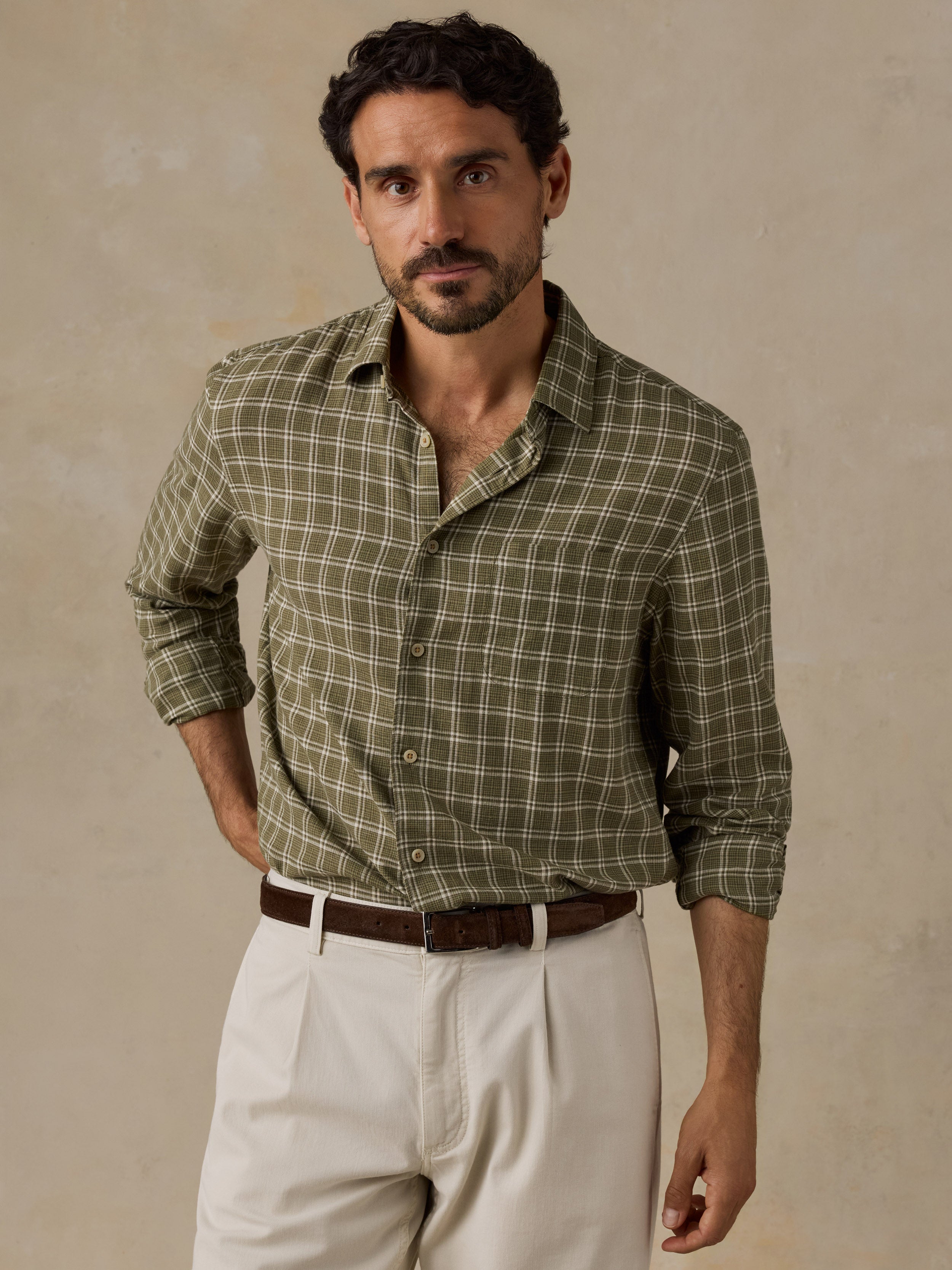 A man wearing a long-sleeve button-up shirt with an olive green and brown plaid pattern, paired with off-white trousers and a brown belt, standing against a textured beige background.