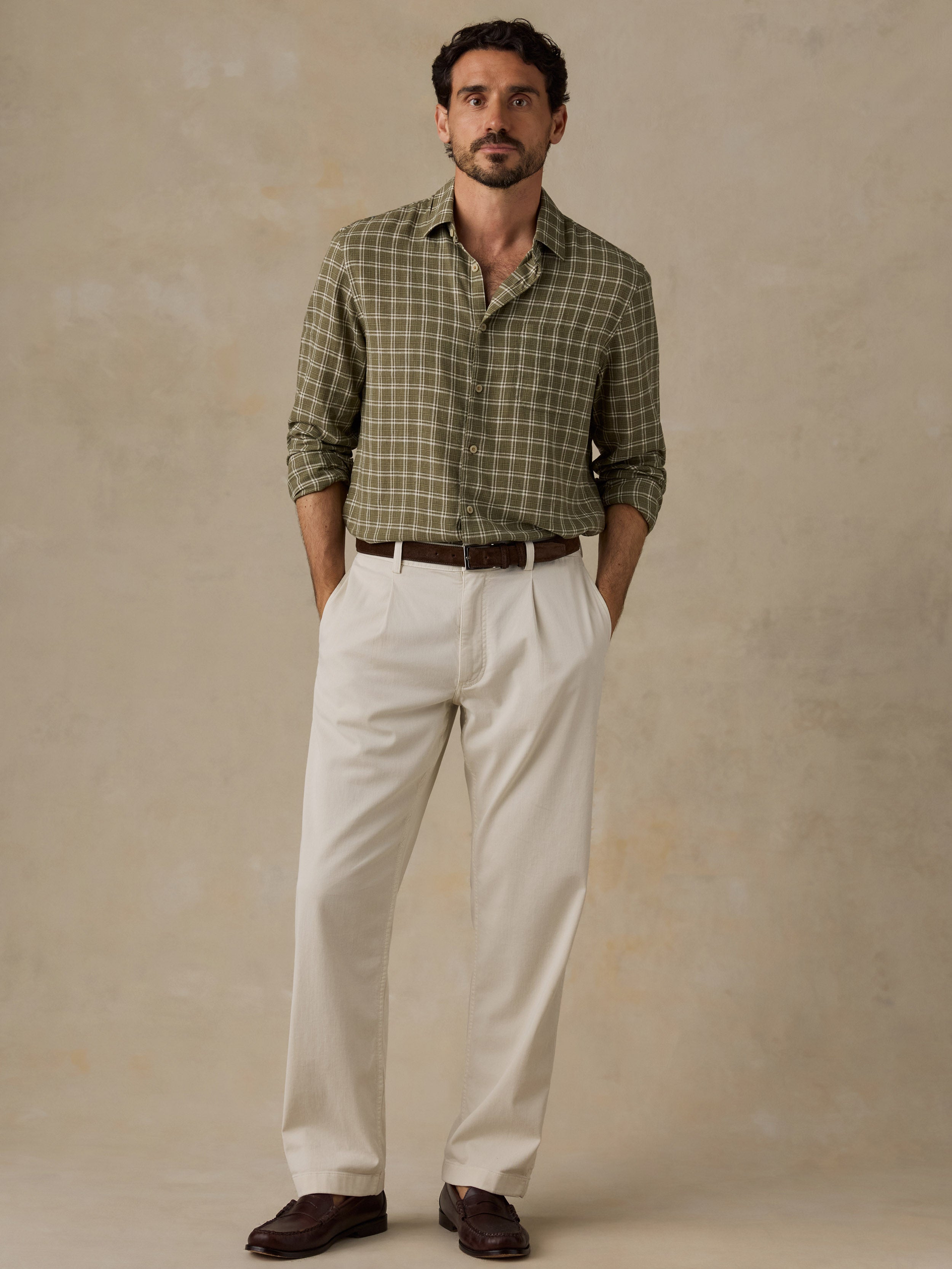 A man wearing a green and beige plaid button-down shirt, off-white trousers, and dark brown loafers, standing against a textured beige studio backdrop.