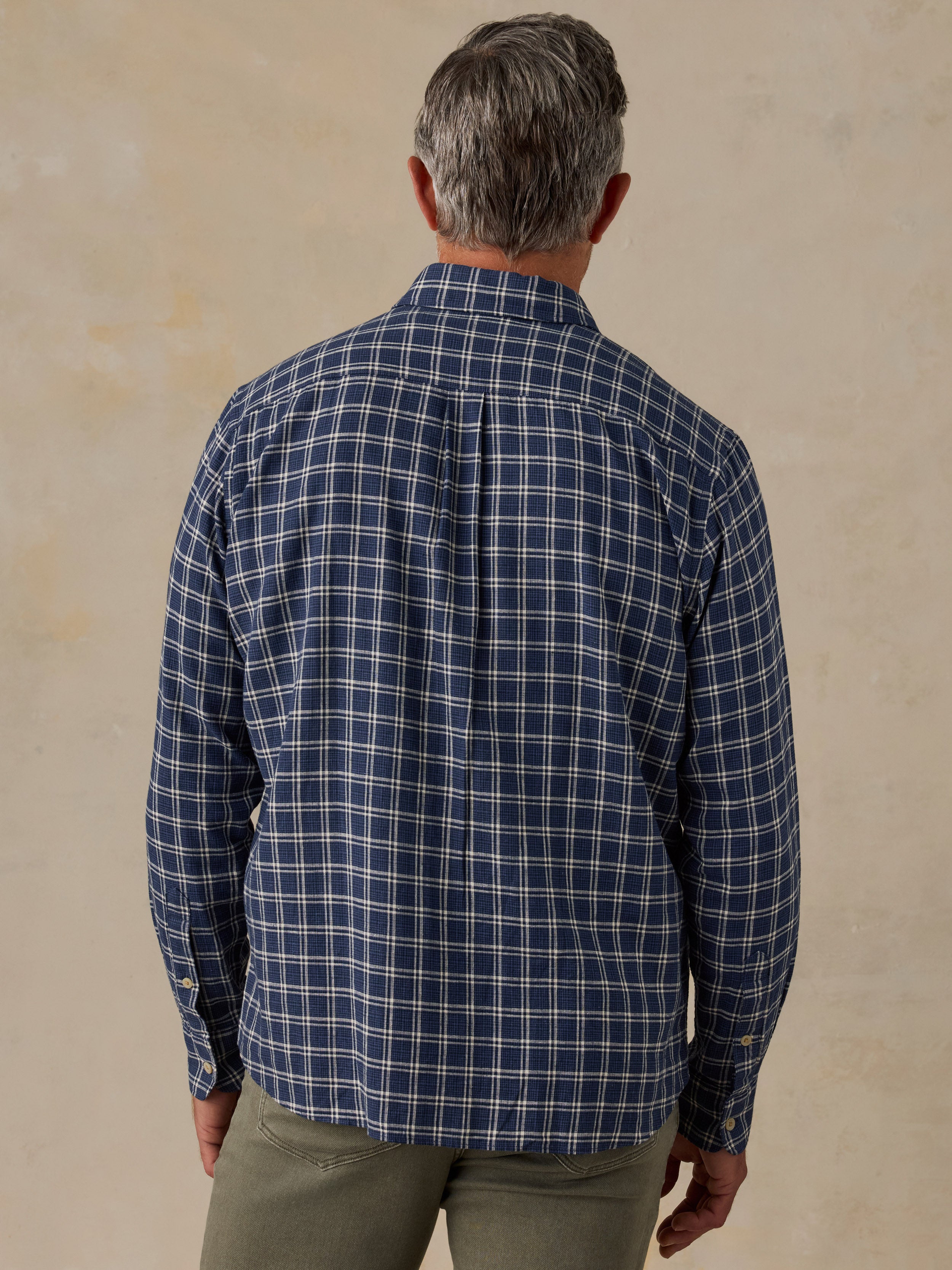 A man viewed from behind wearing a navy blue plaid button-down shirt with white and light blue grid lines, paired with olive green pants, against a textured beige background.