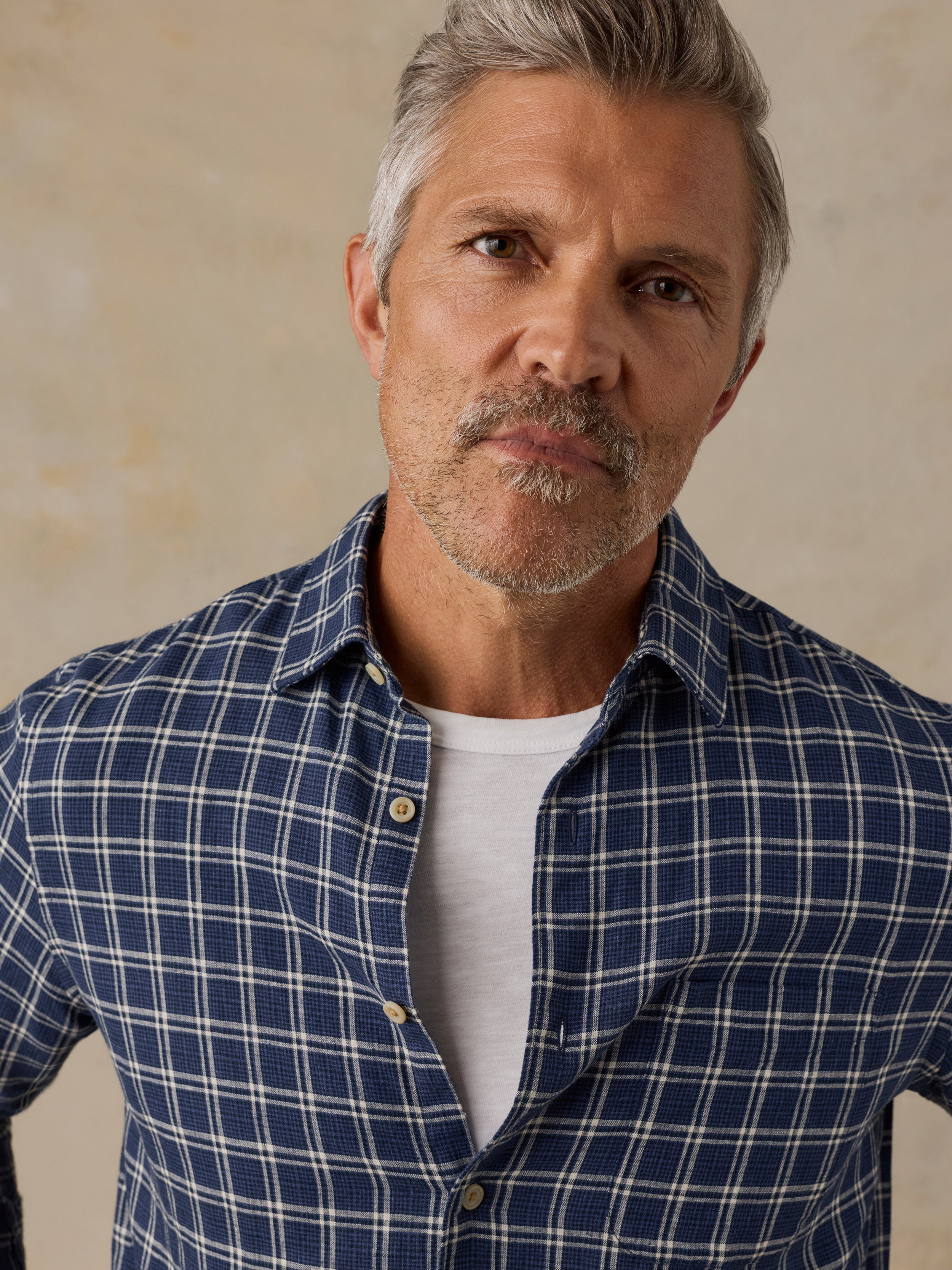 A close-up portrait of a man with gray hair and a beard wearing a navy blue and white plaid button-down shirt over a white t-shirt, set against a textured beige background.