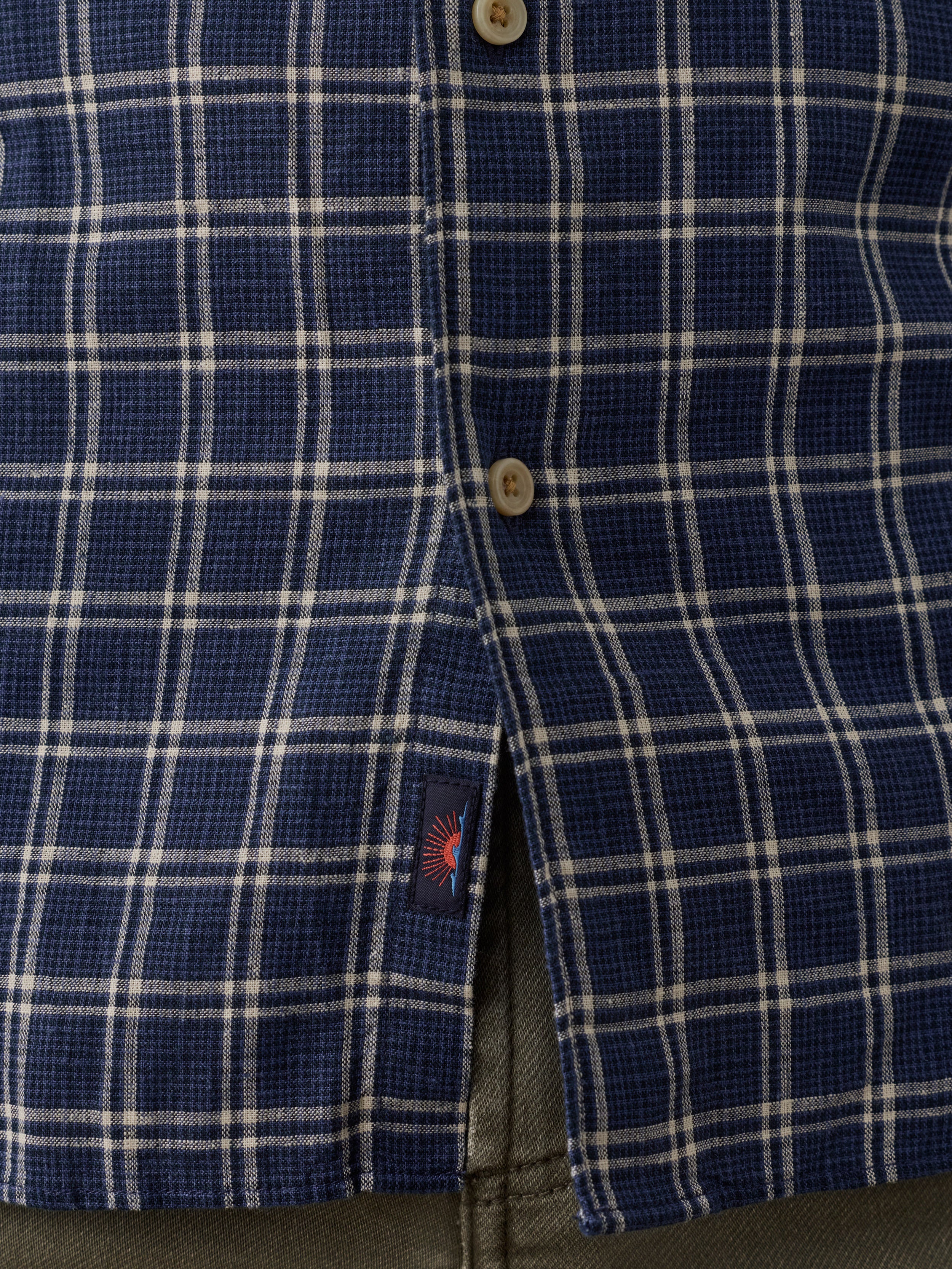Close-up of a blue and white plaid button-down shirt with a small Faherty brand label near the bottom hem.