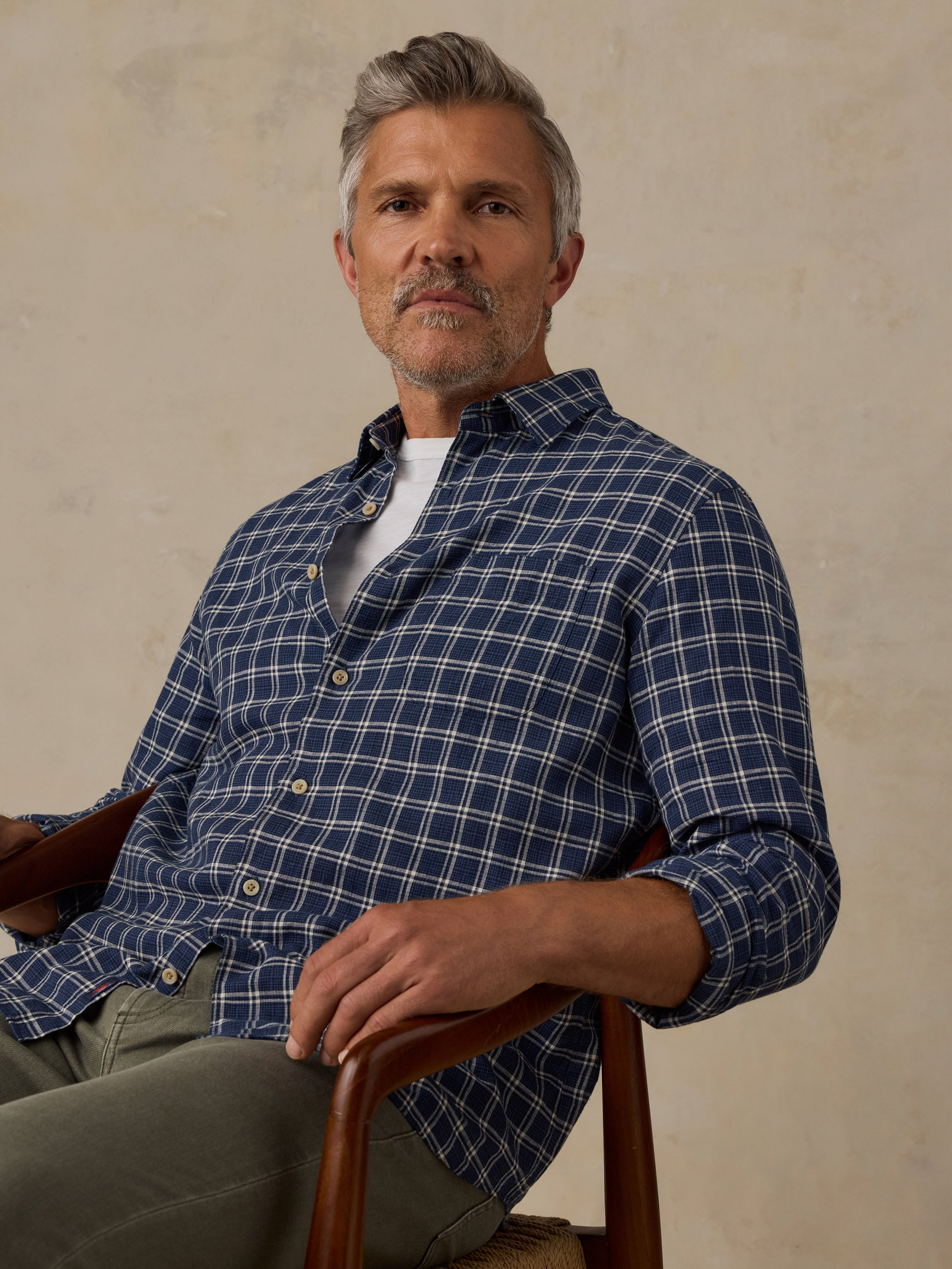 A man with gray hair and a beard sits in a wooden chair wearing a navy blue and white plaid button-down shirt over a white undershirt, paired with olive green trousers, against a textured beige wall.
