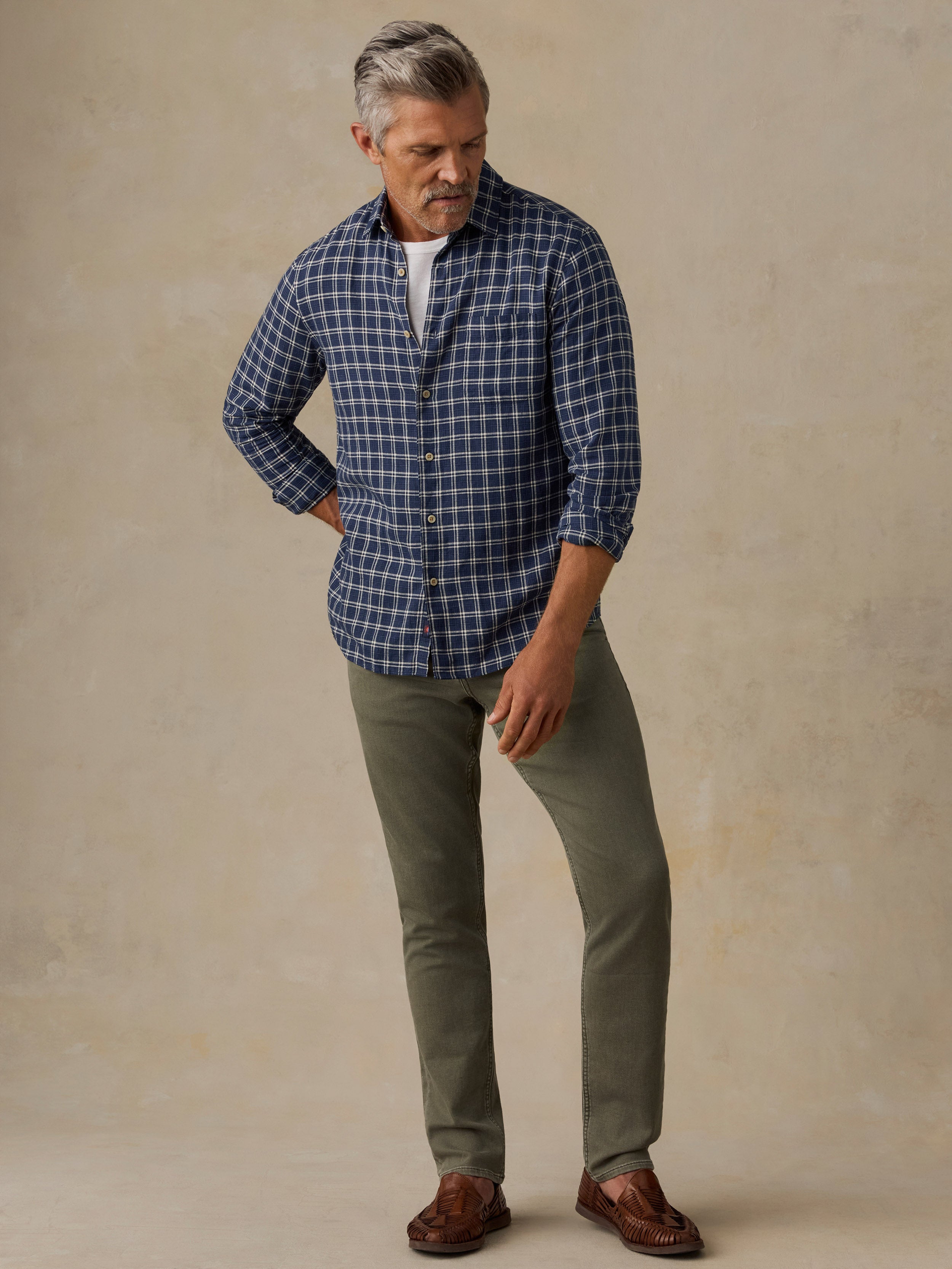 A man wearing a blue and white plaid button-down shirt, olive green pants, and brown leather loafers, standing against a textured beige studio backdrop.