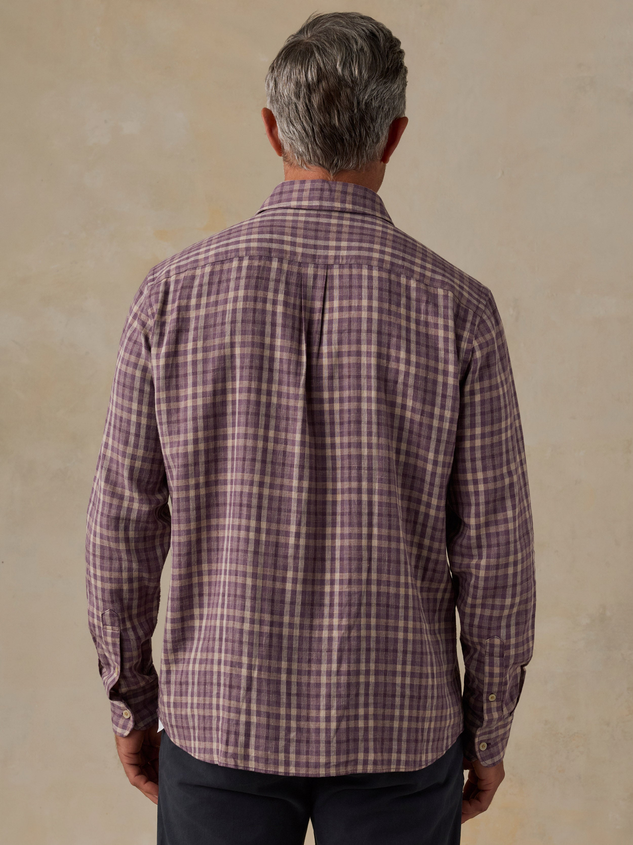 A man viewed from behind wearing a long-sleeve plaid button-down shirt in a muted purple and beige pattern, paired with dark trousers, against a textured beige background.