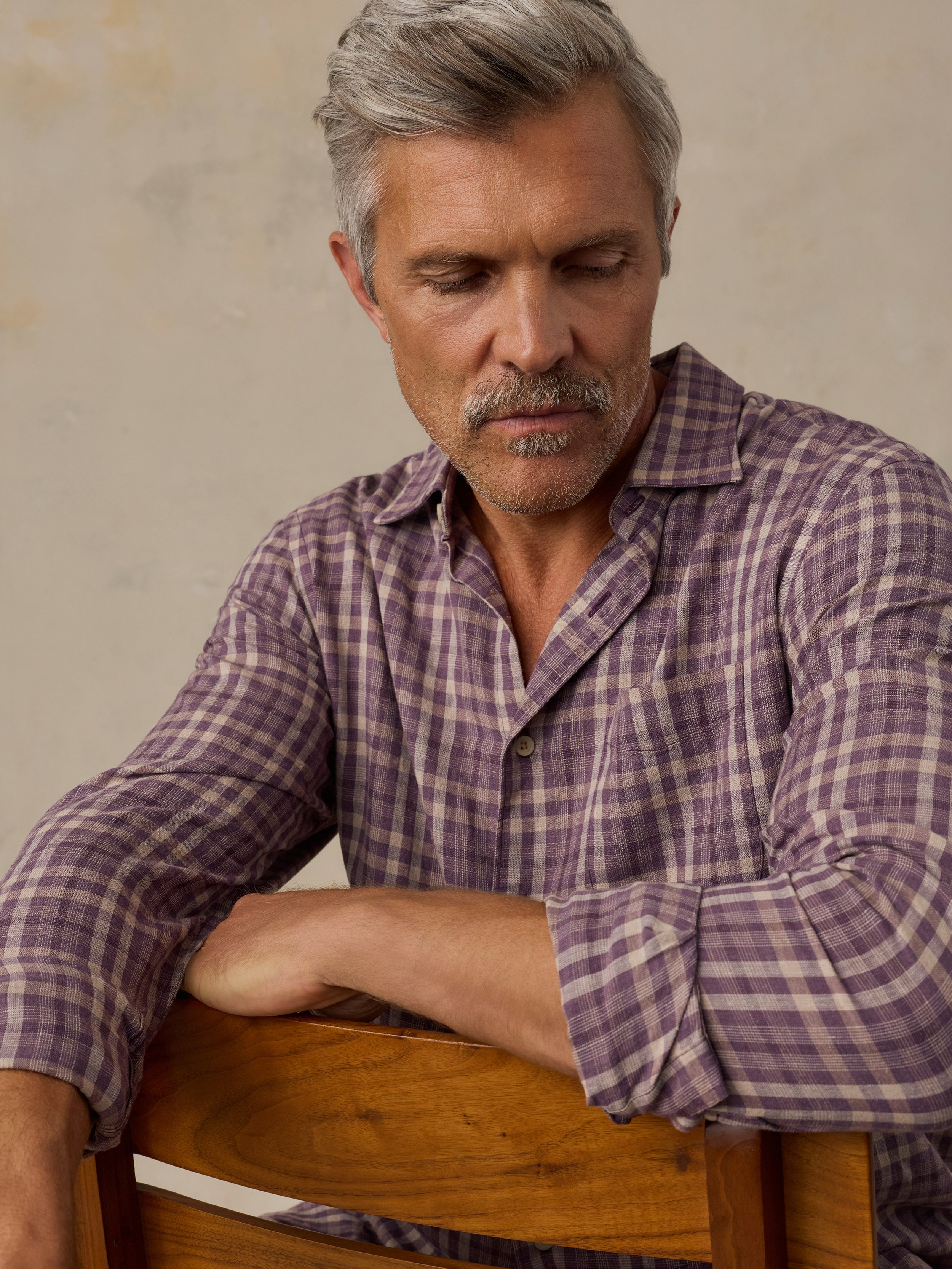 A man with gray hair and a mustache wearing a purple and beige plaid button-down shirt, leaning on a wooden chair against a textured beige wall.