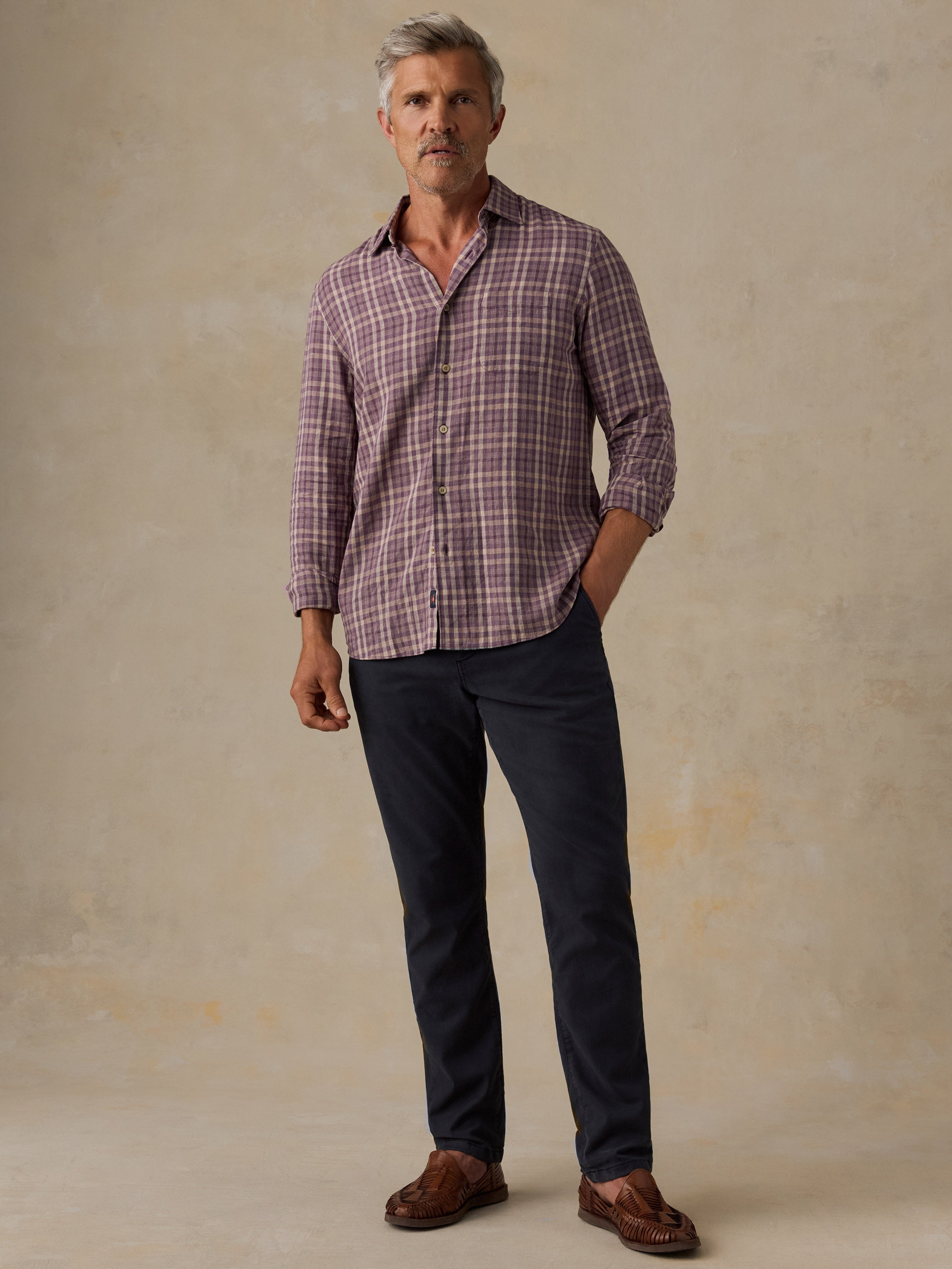 A man wearing a long-sleeve plaid button-down shirt in muted purple and white, paired with dark navy trousers and brown loafers, standing against a textured beige studio backdrop.