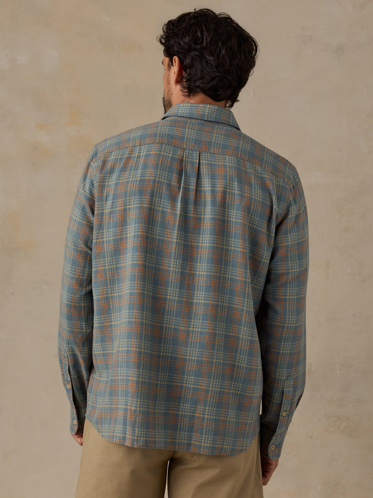 A man viewed from behind wearing a long-sleeve plaid button-up shirt in muted blue-gray, beige, and brown tones, paired with khaki pants, against a textured beige wall., image 5 of 6