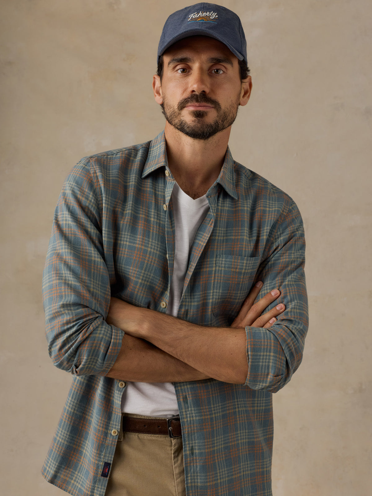 A man wearing a blue and green plaid button-down shirt over a white t-shirt, khaki pants, and a blue baseball cap with 'Faherty' embroidered on the front, standing with arms crossed against a textured beige wall., image 2 of 6