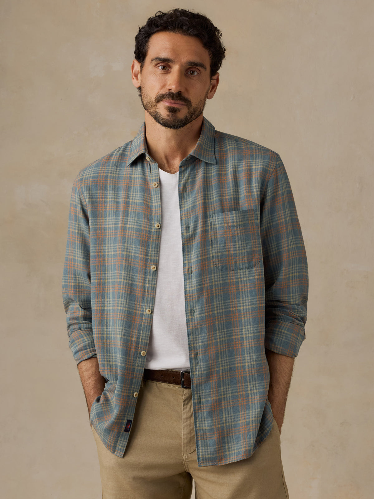 A man wearing a muted blue-green plaid button-down shirt over a white t-shirt and khaki pants, standing against a textured beige wall., image 1 of 6