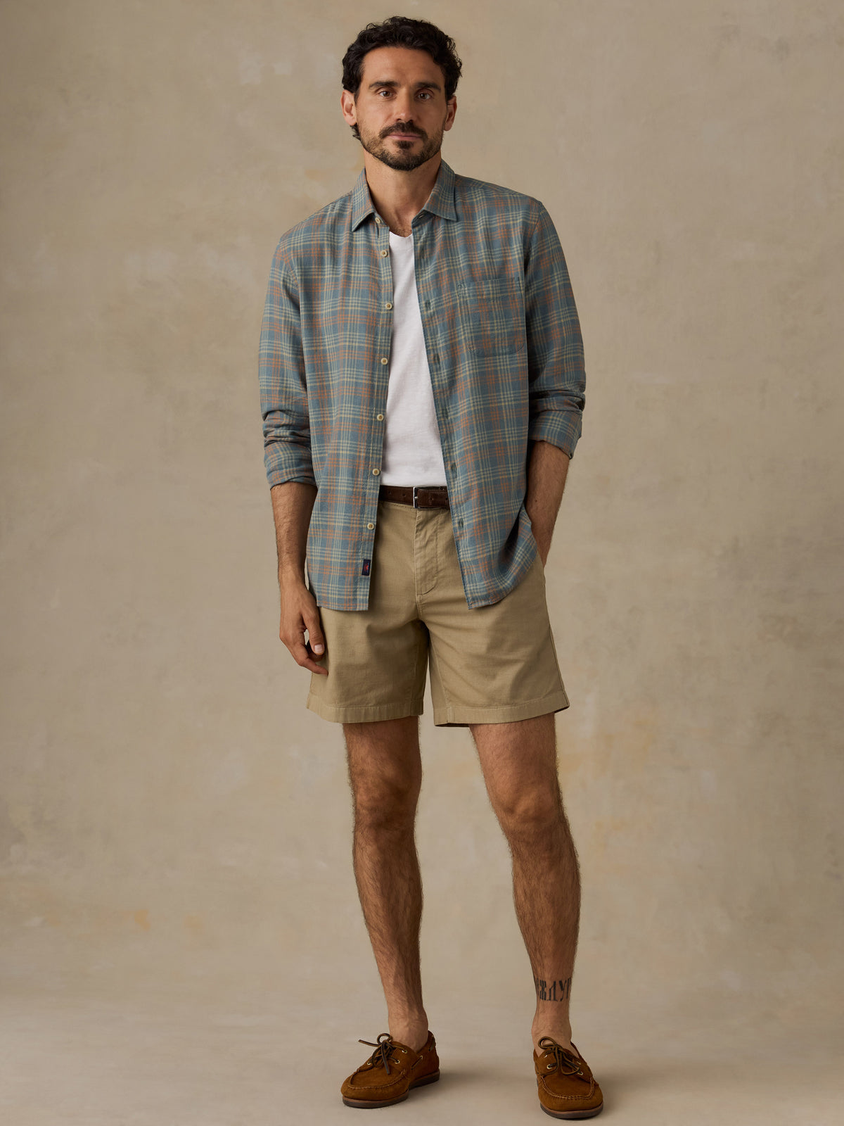 A man modeling a long-sleeve plaid button-down shirt in muted blue and green tones over a white t-shirt, paired with khaki shorts and brown suede shoes, against a textured beige studio backdrop., image 6 of 6