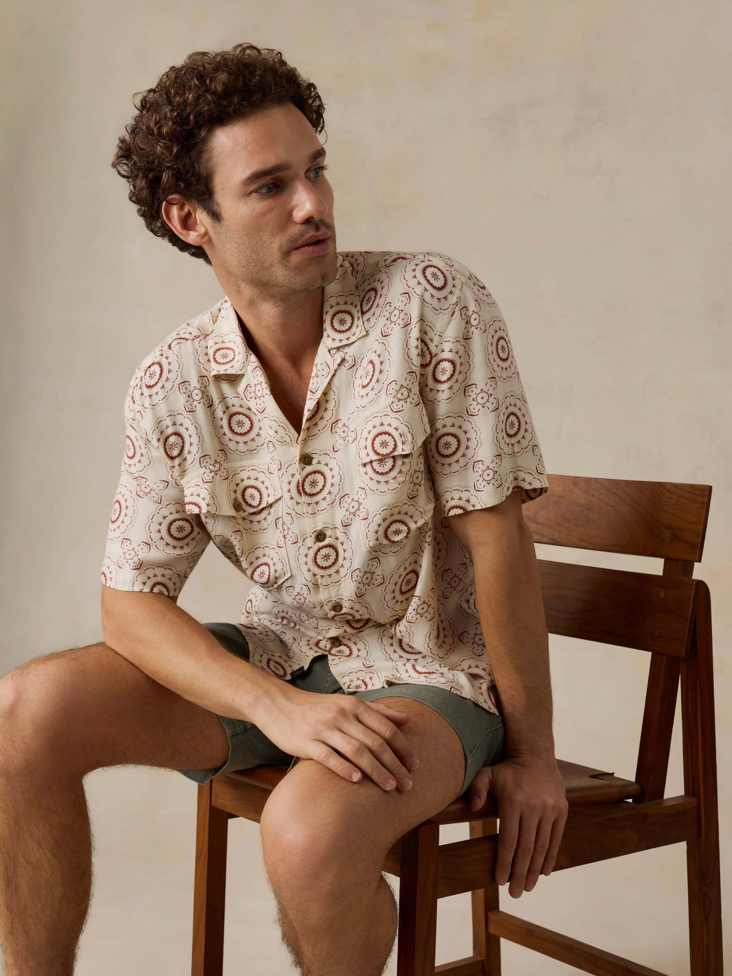 Man sitting on a wooden chair wearing a patterned shirt and green shorts against a beige background