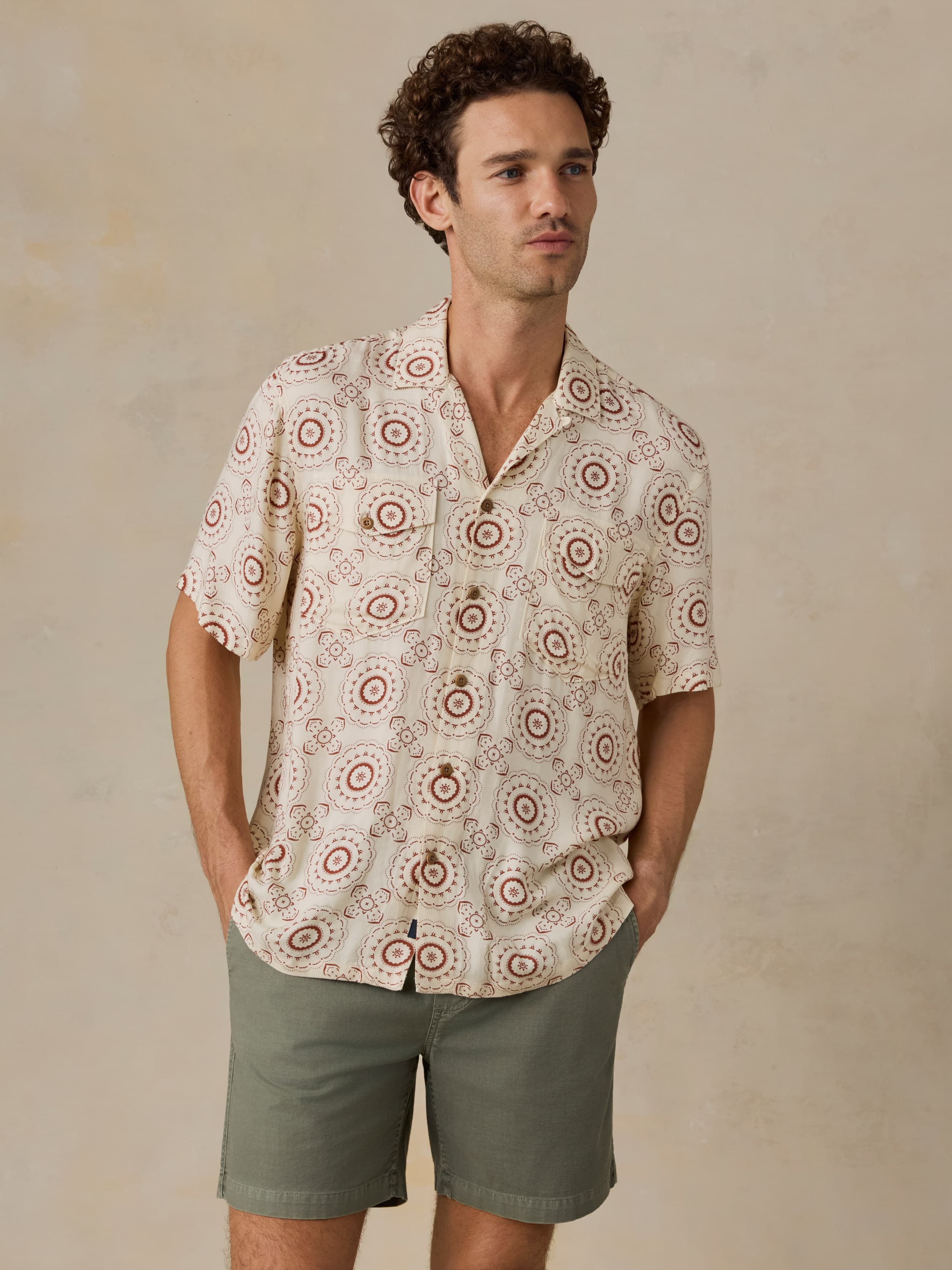 Man wearing a patterned shirt and green shorts against a beige background
