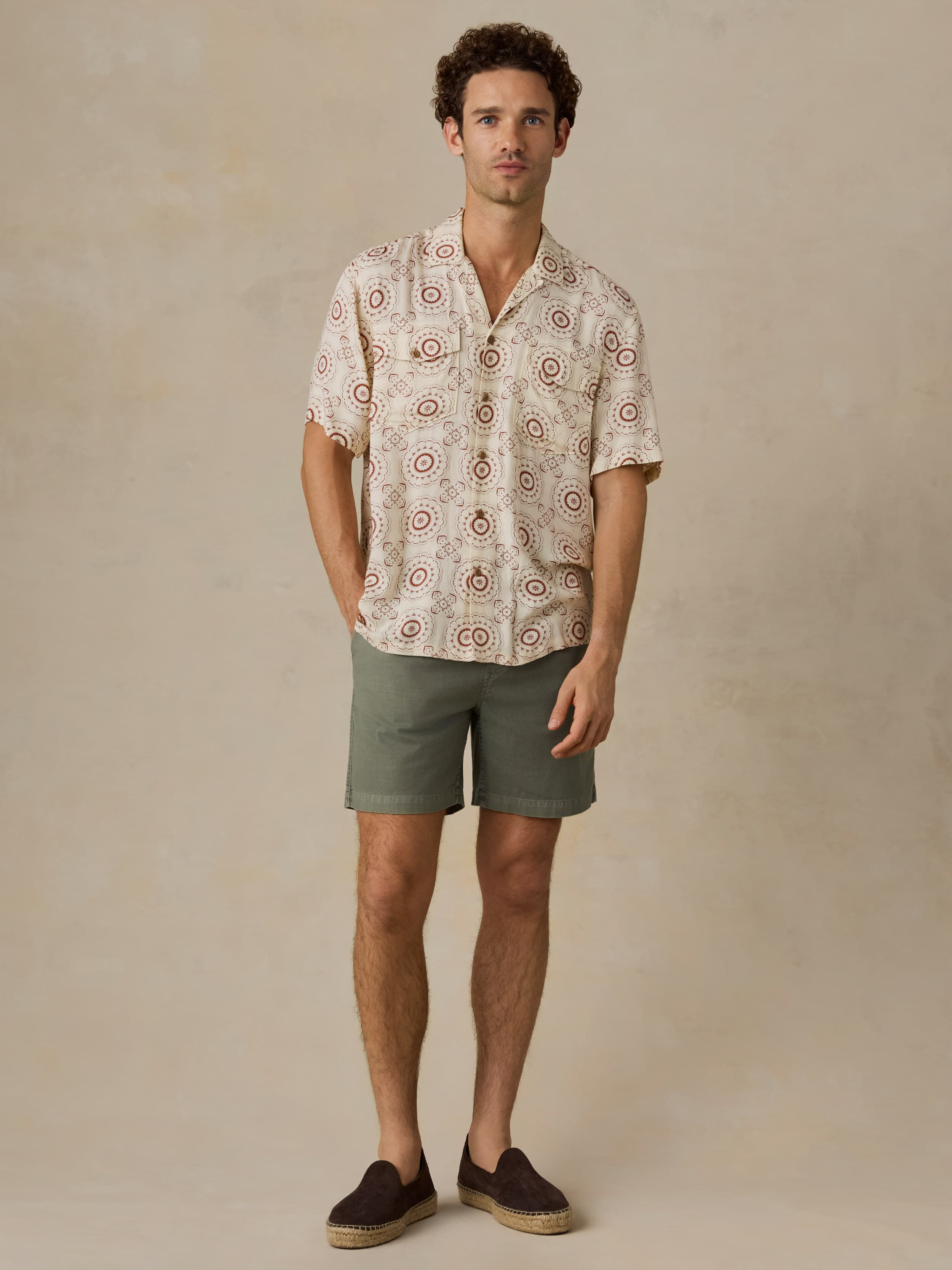Man wearing a patterned shirt and green shorts against a beige background