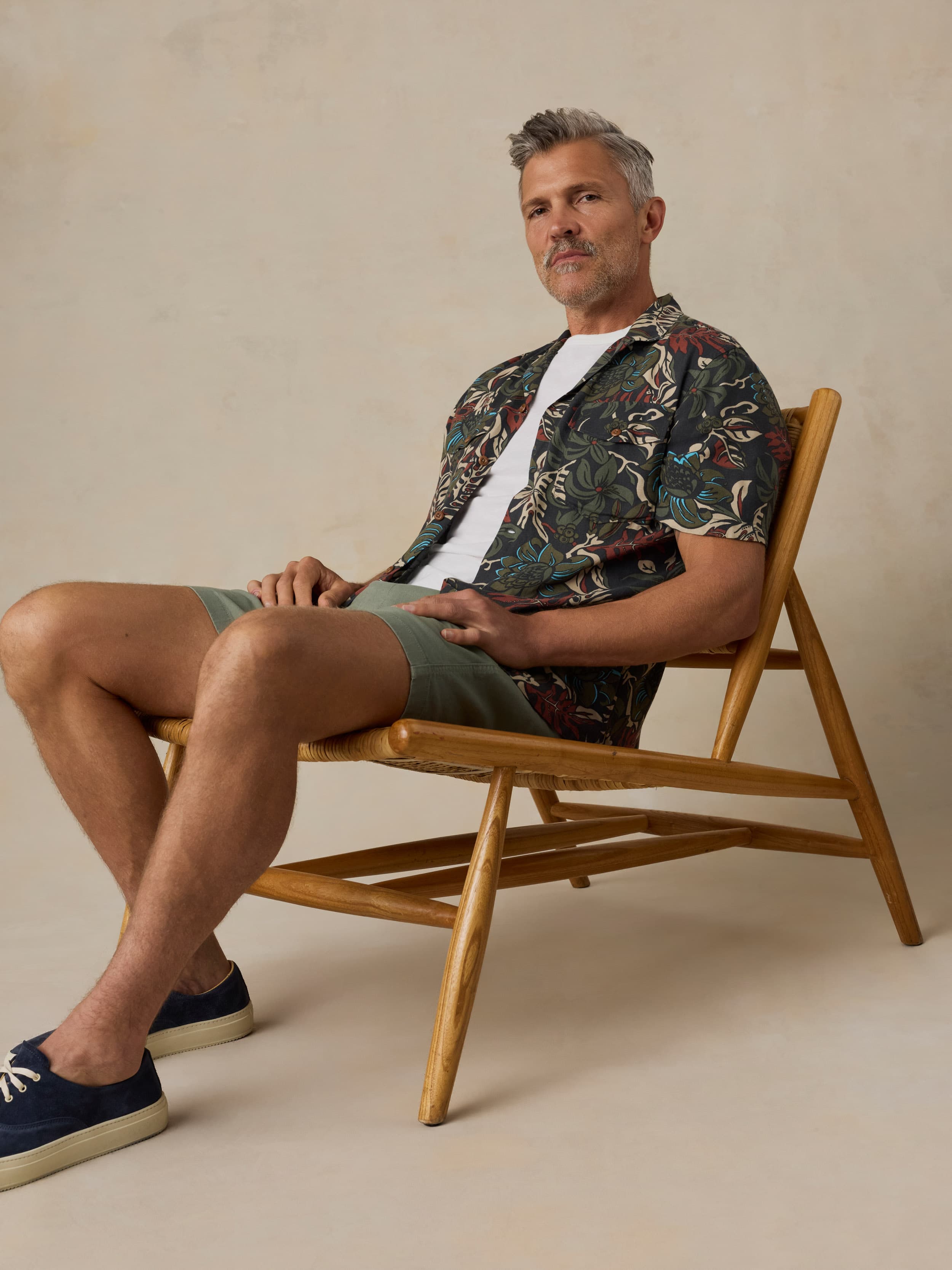 Man sitting on a wooden chair wearing a tropical patterned shirt and green shorts against a beige background