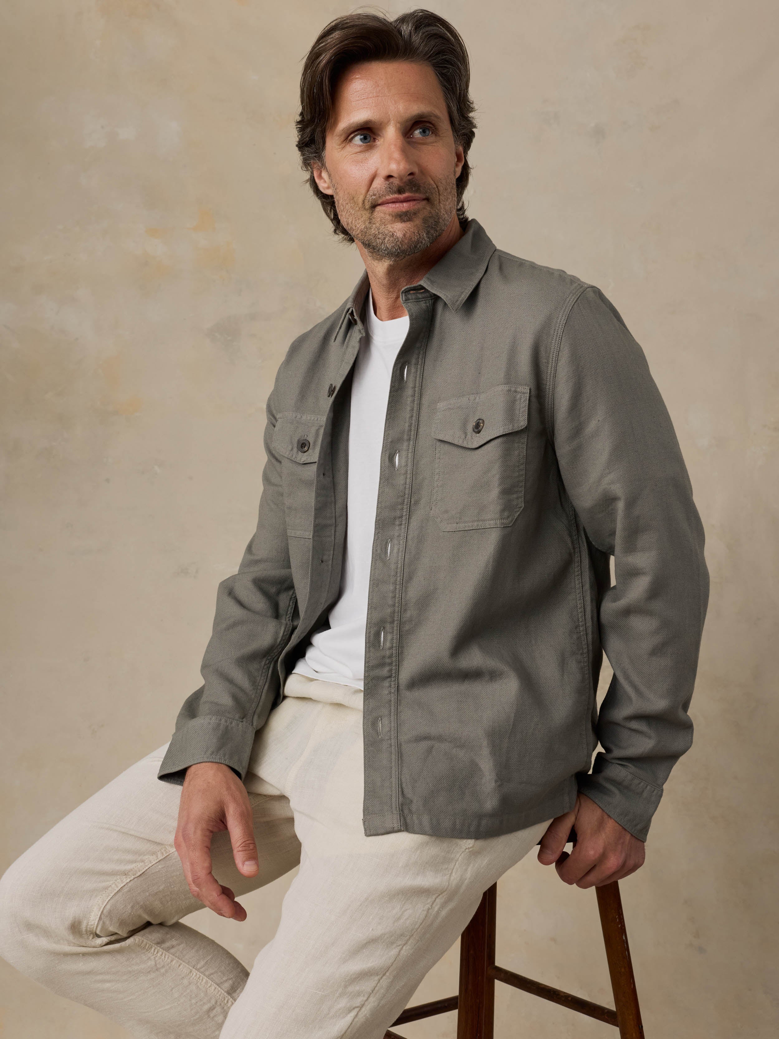 Man sitting on a wooden stool wearing an olive green jacket over a white shirt with beige pants against a beige background