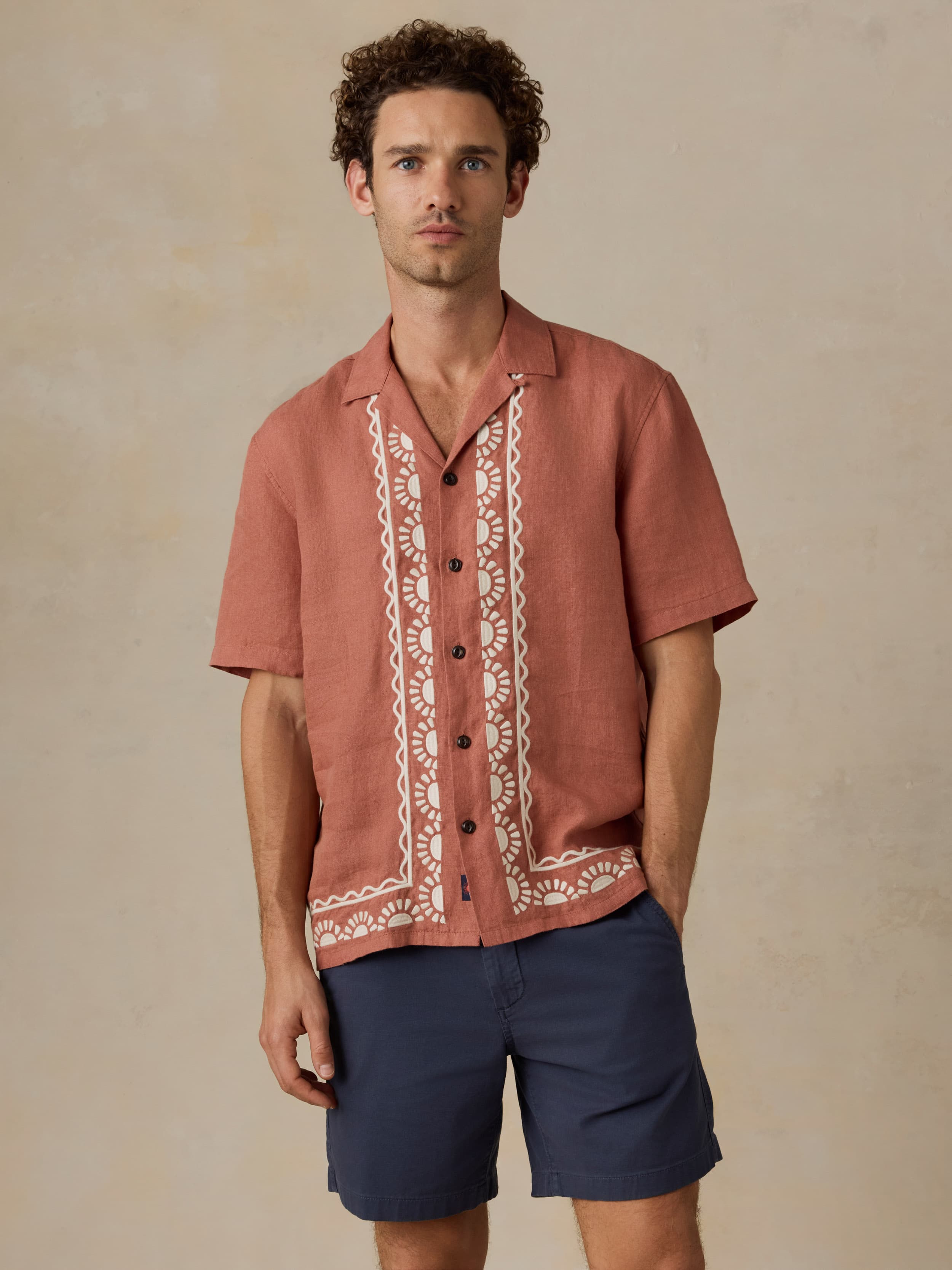 Man wearing a red patterned embroidered shirt and navy shorts against a beige background