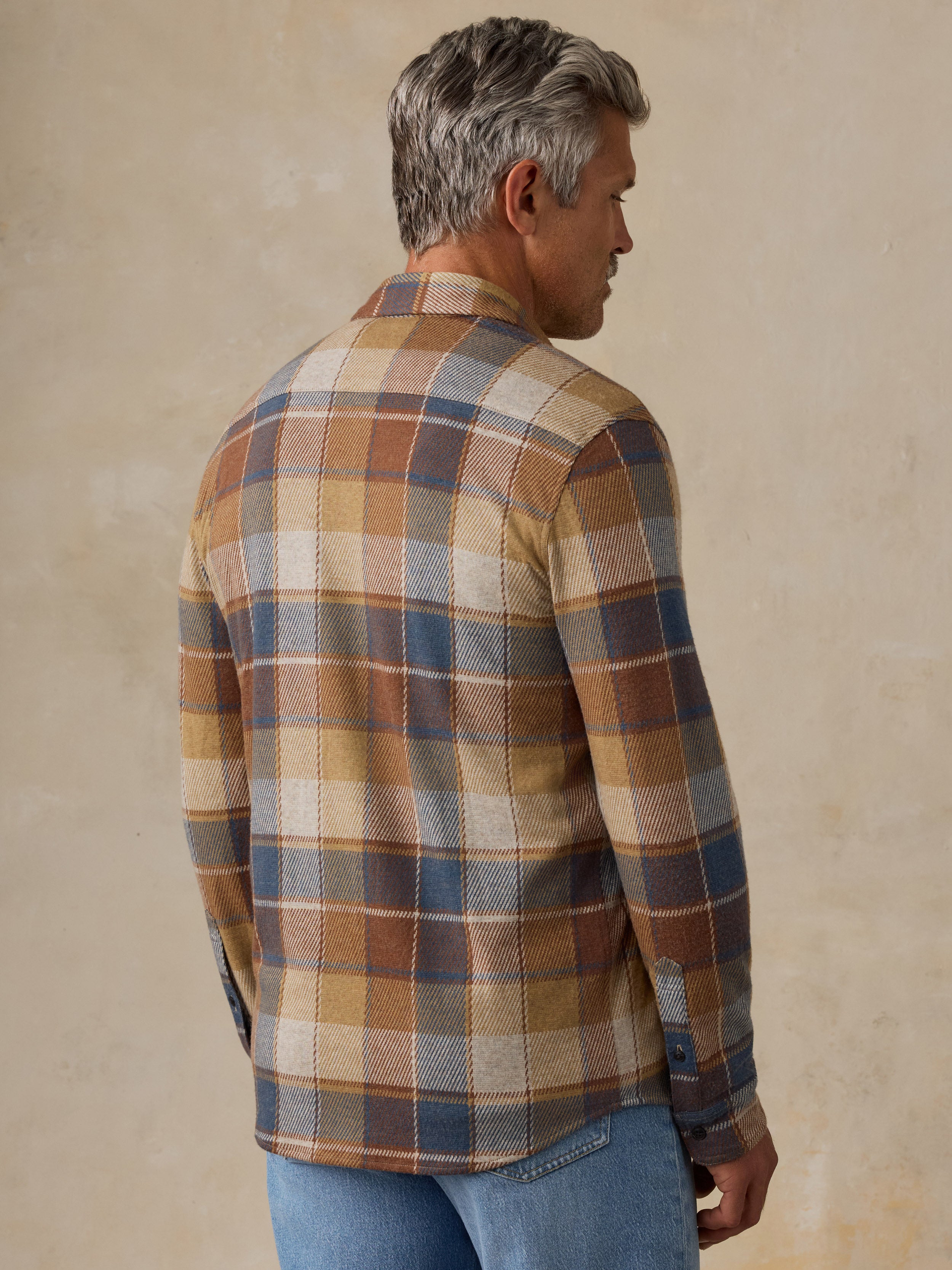 A man wearing a long-sleeve plaid button-down shirt, shown from a three-quarters back view. The shirt features a multi-colored plaid pattern with brown, beige, blue, and white stripes.