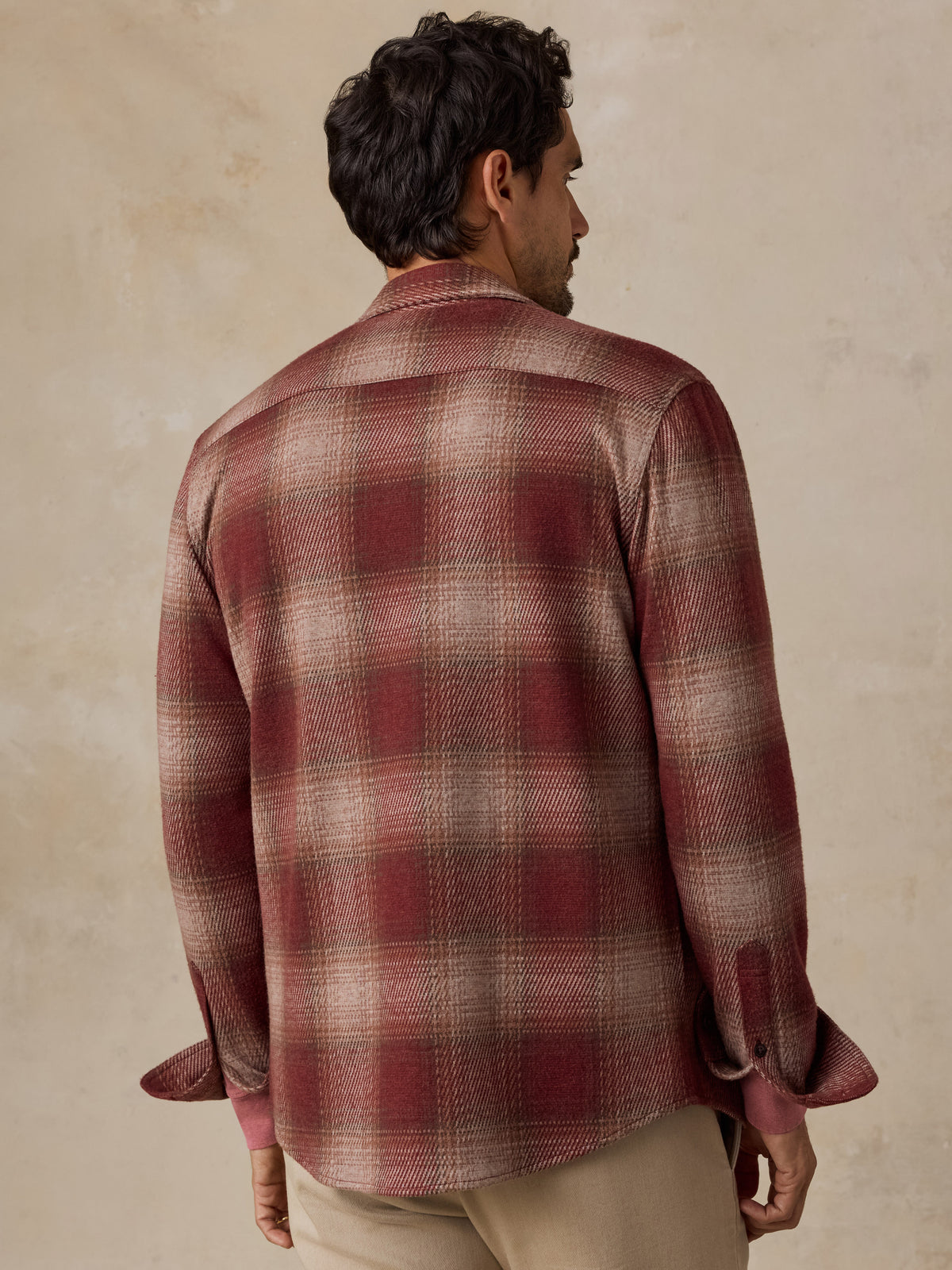 A man viewed from behind wearing a long-sleeve plaid button-up shirt in muted red and beige tones, paired with khaki pants, against a textured beige studio backdrop., image 5 of 7