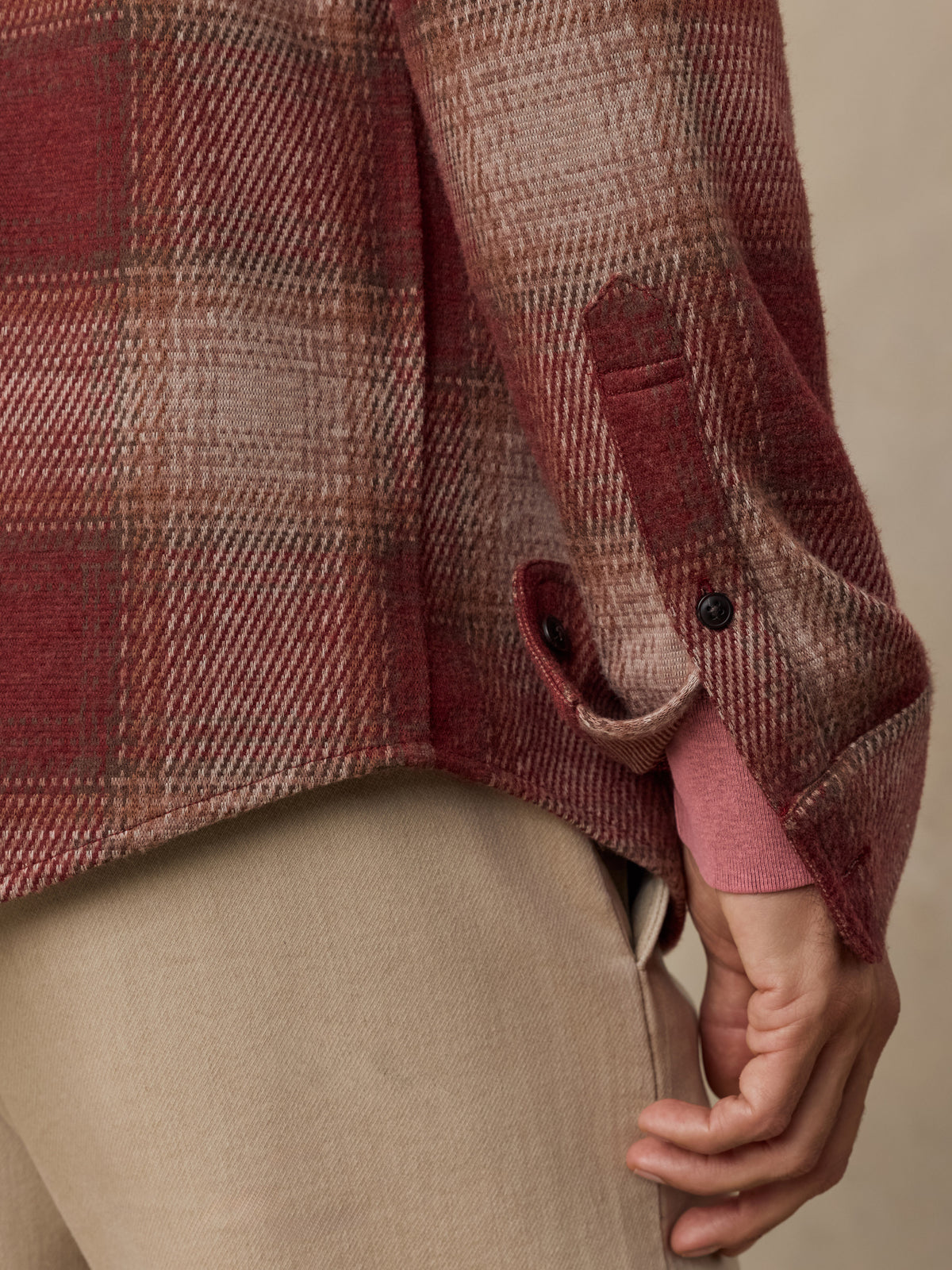Close-up of a man wearing a muted red and beige plaid overshirt with a button cuff and beige trousers, against a plain beige background., image 4 of 7