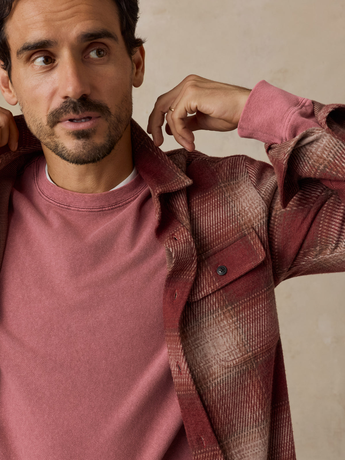 A man wearing a dusty rose crewneck sweatshirt layered under an open rust-brown plaid flannel shirt, posed against a neutral textured beige background., image 6 of 7