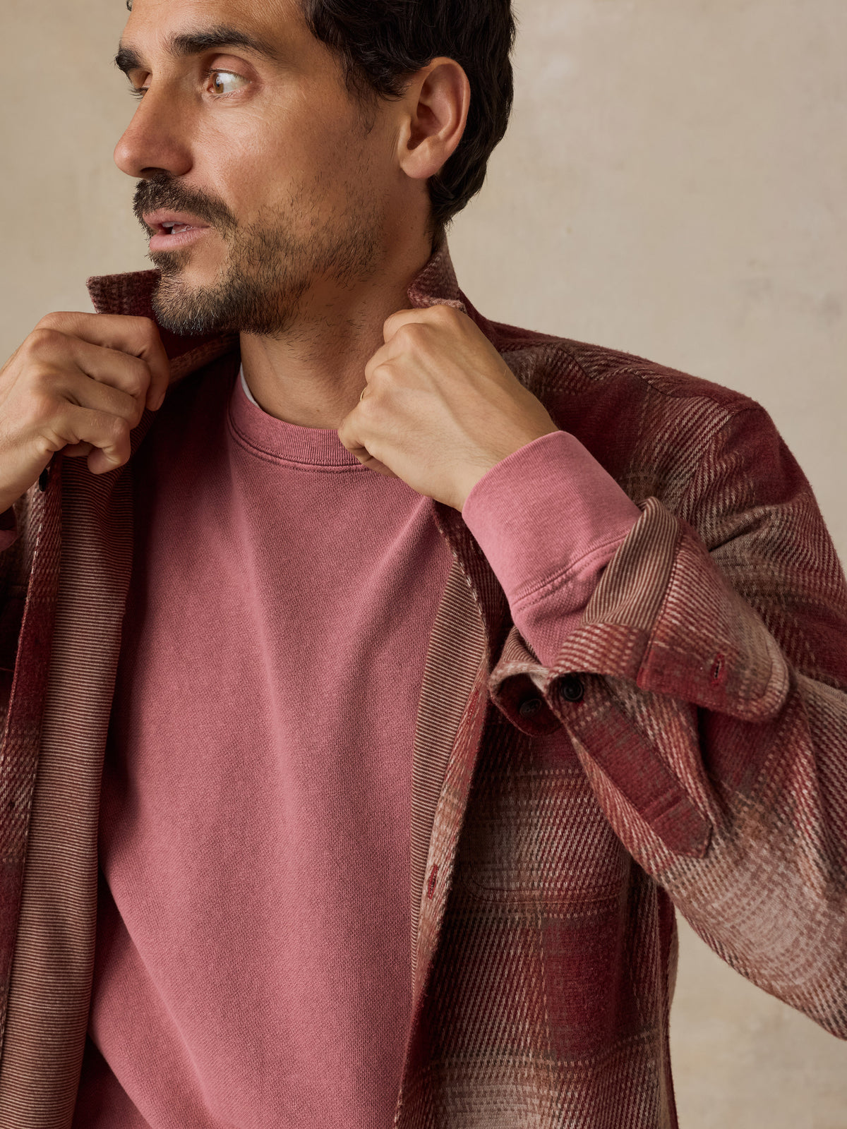 A man wearing a dusty rose crewneck sweatshirt under an open rust-brown plaid overshirt, posing against a neutral beige background., image 3 of 7