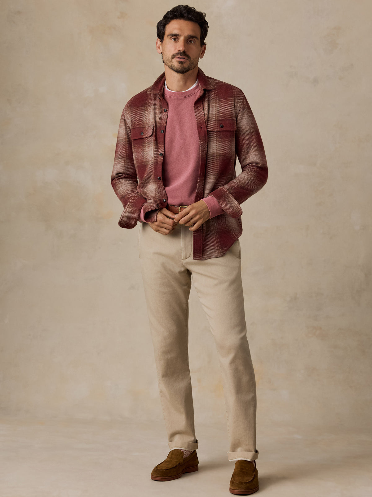 A man stands against a textured beige studio backdrop wearing a burgundy and brown plaid flannel shirt over a dusty pink crewneck sweater, paired with beige trousers and brown suede loafers., image 7 of 7