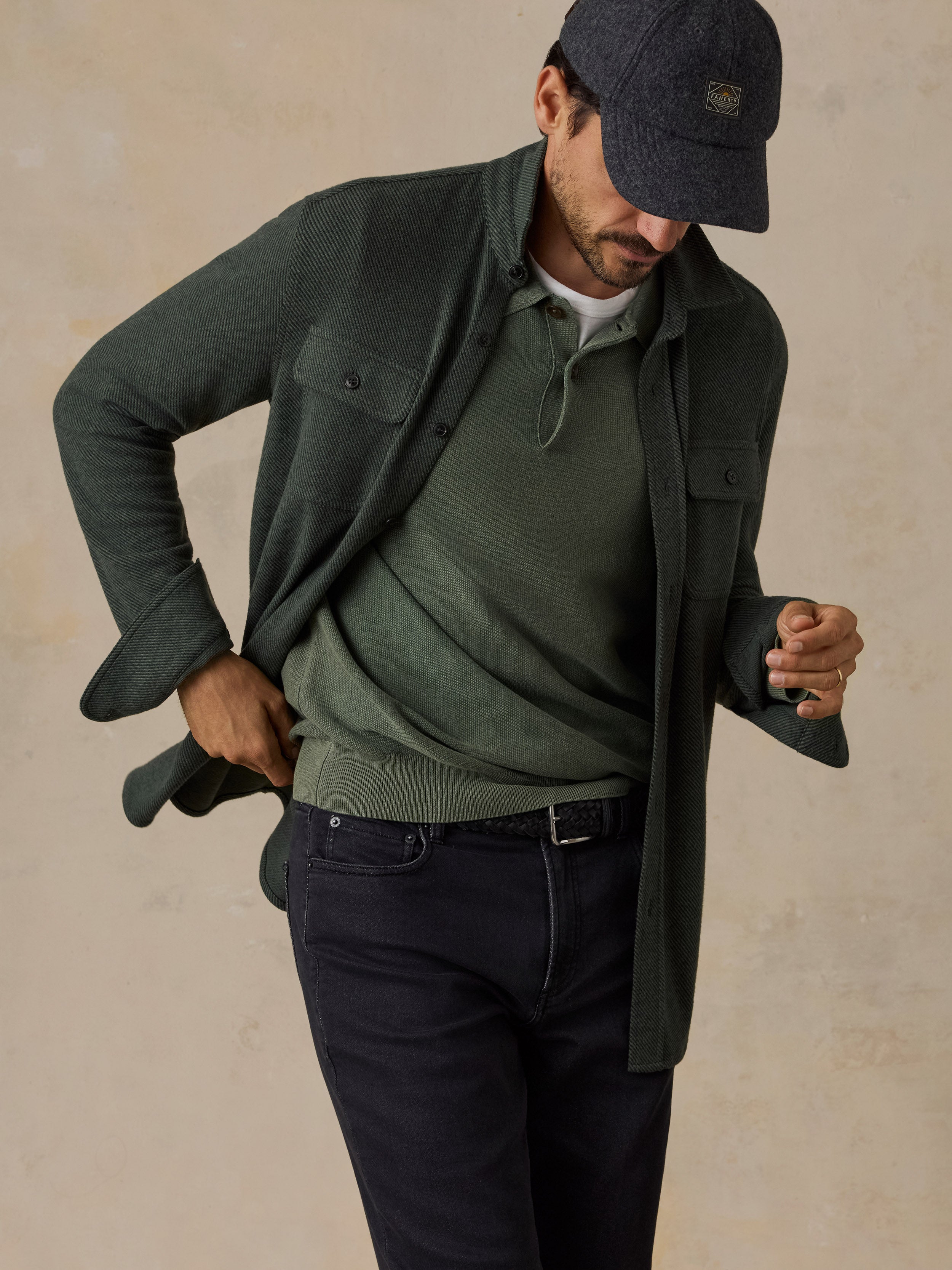 A man wearing a dark green button-up overshirt over an olive green knit sweater, paired with dark navy jeans and a dark baseball cap, posing against a textured beige background.