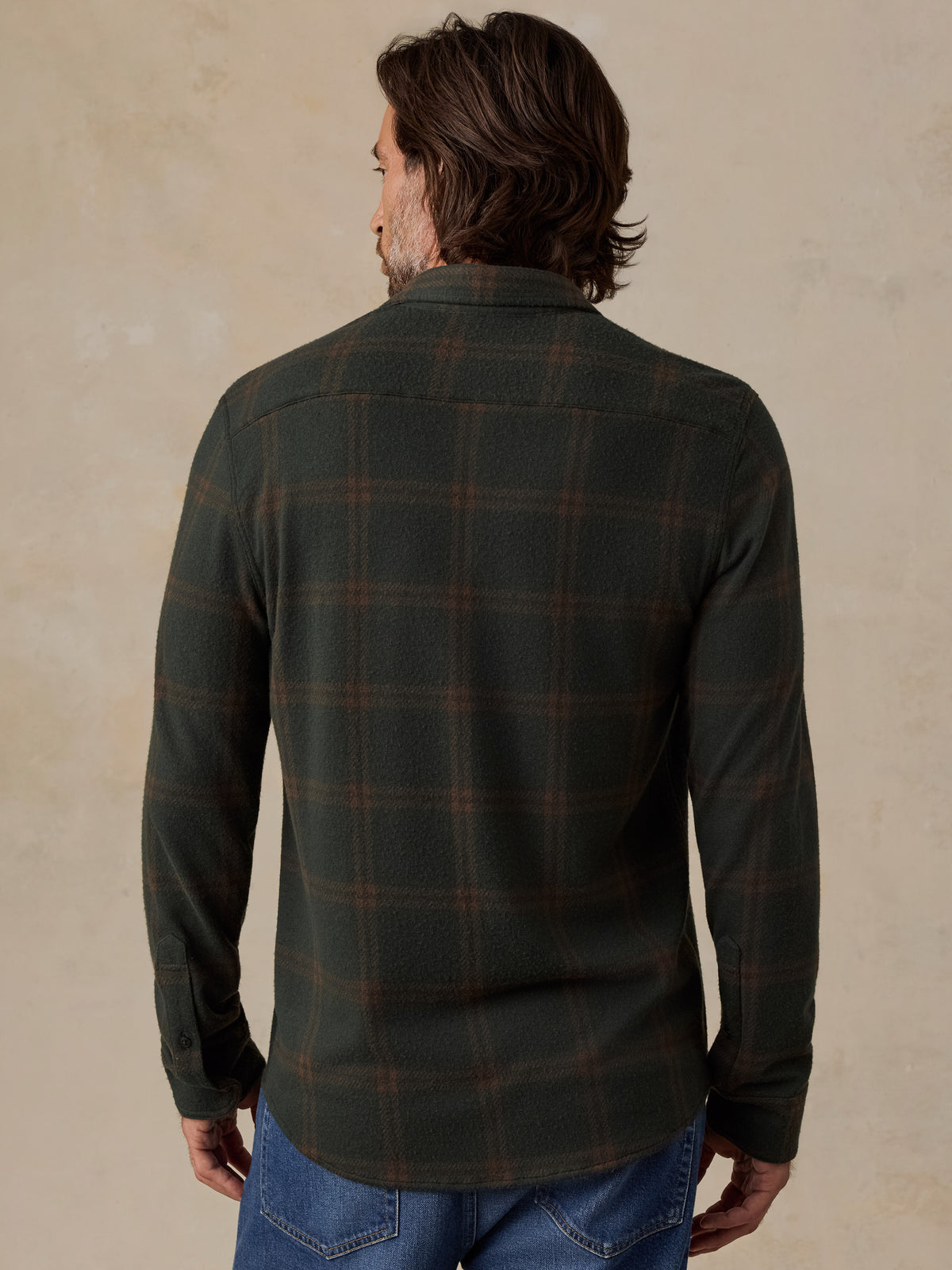 A man viewed from the back wearing a dark green long-sleeve plaid flannel shirt and blue jeans, standing against a textured beige wall., image 3 of 5