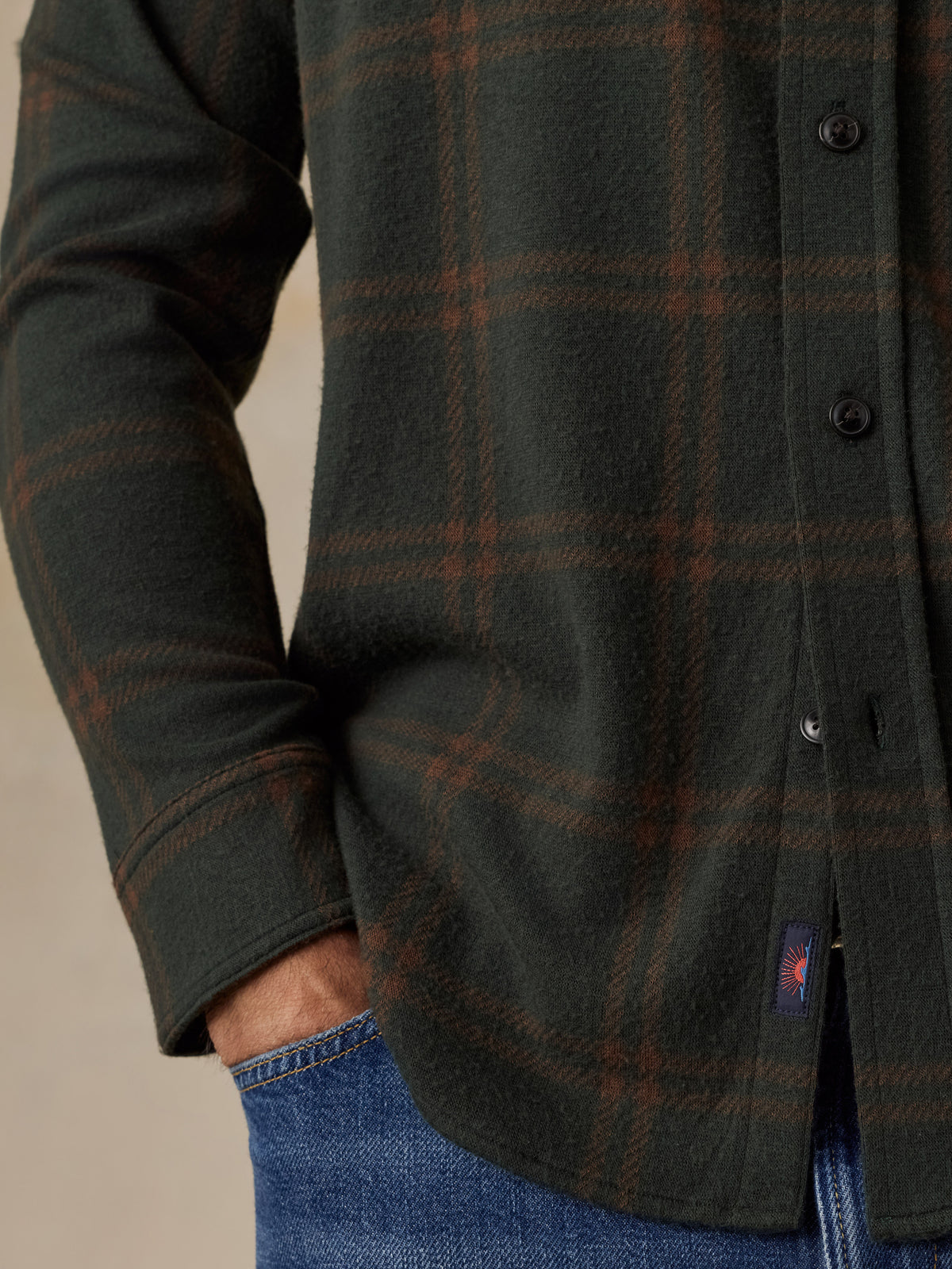 Close-up of a man wearing a dark olive green and brown plaid button-up shirt with blue jeans, showing the fabric texture and a small red and blue Faherty brand tab near the hem., image 4 of 5