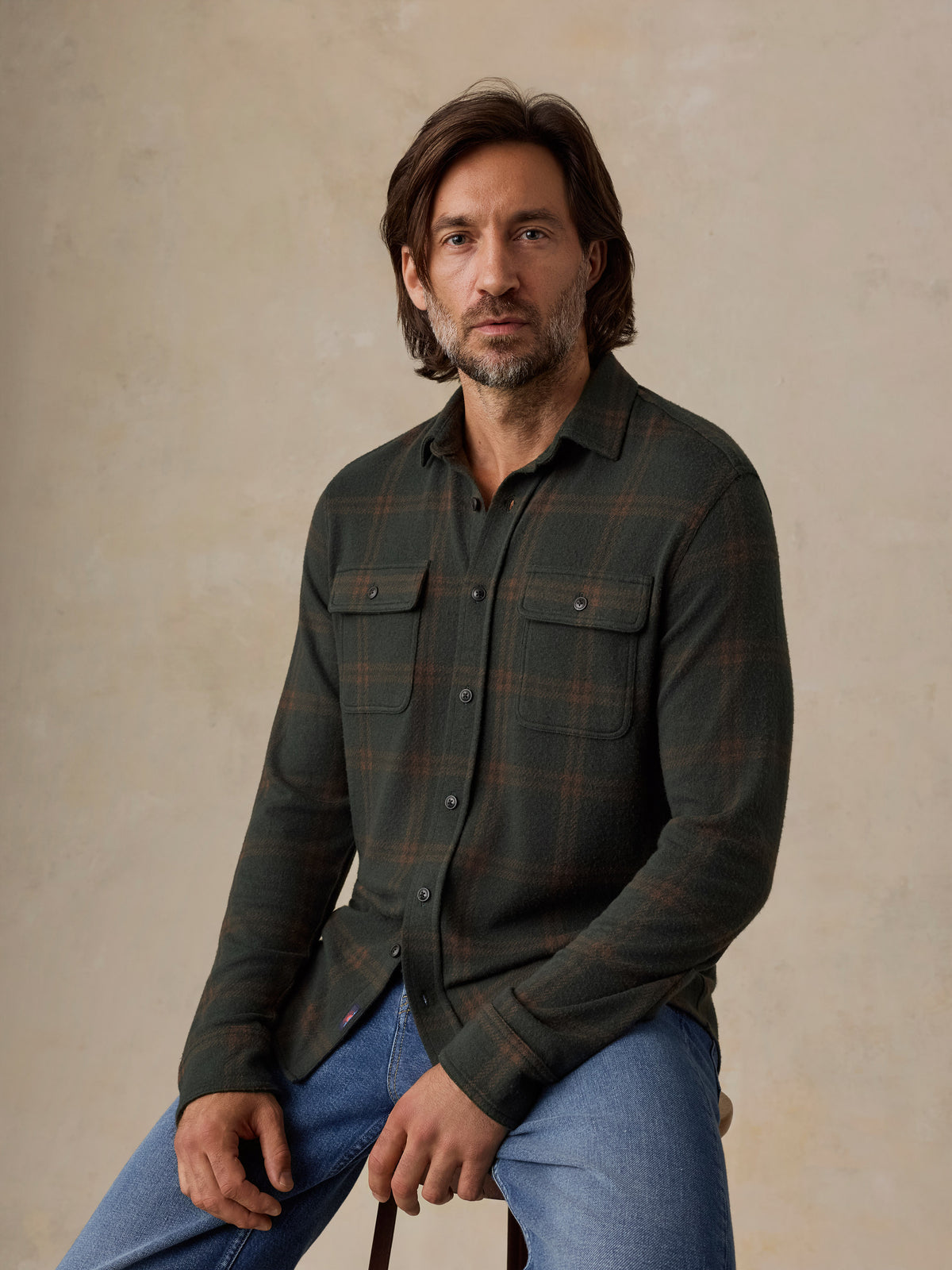 A man wearing a dark green plaid button-down shirt and blue jeans, sitting against a textured beige studio backdrop., image 5 of 5