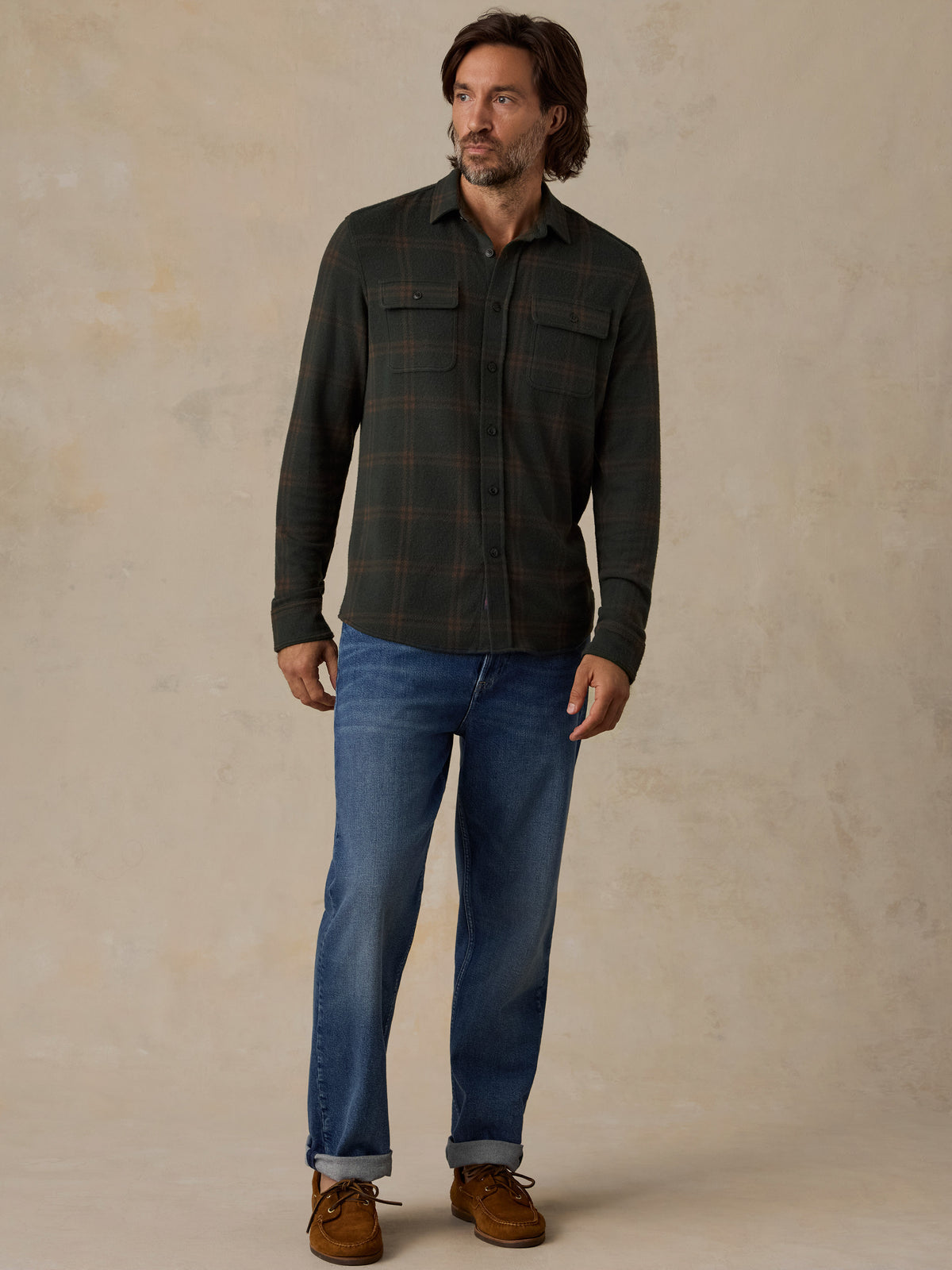 A man wearing a dark green and brown plaid button-down flannel shirt, blue jeans, and brown suede boots, standing against a textured beige studio backdrop., image 2 of 5