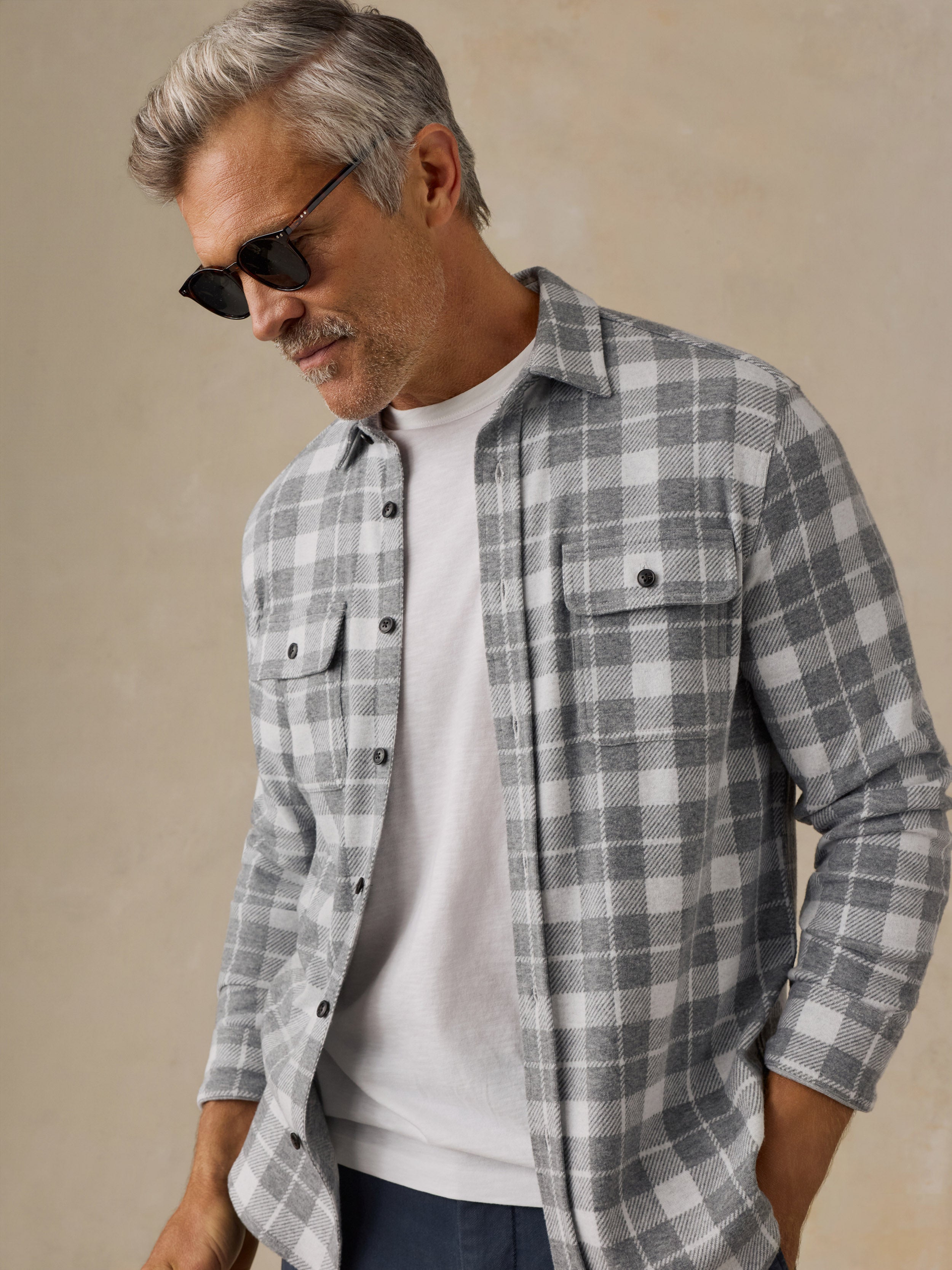 A man wearing a gray and white plaid button-down flannel shirt over a white t-shirt, styled with dark sunglasses and navy pants, set against a neutral beige background.