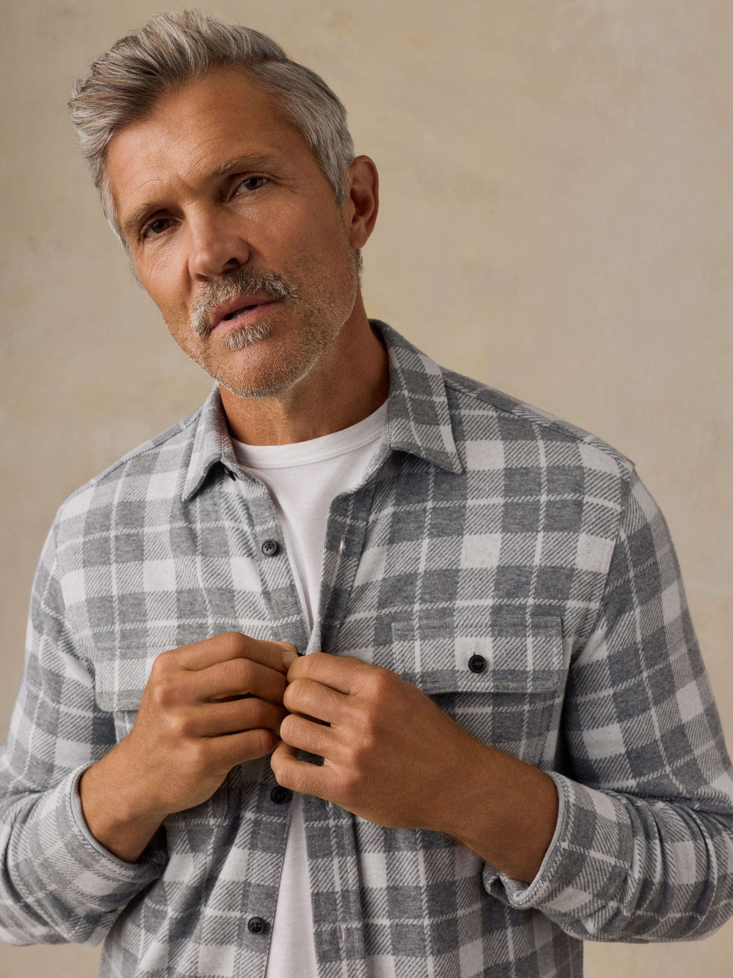 A man with gray hair and a beard wearing a gray and white plaid button-down shirt over a white t-shirt, adjusting the collar buttons against a textured beige background.