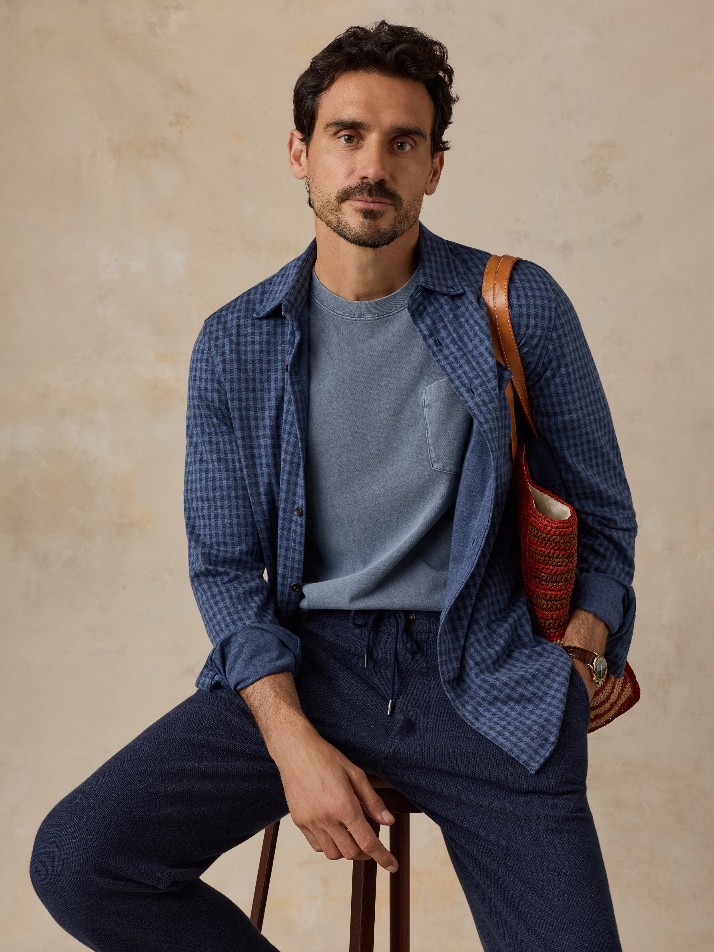 A man wearing a blue gingham overshirt over a slate blue t-shirt and navy drawstring trousers, sitting on a stool against a textured beige wall and holding a brown leather shoulder bag.